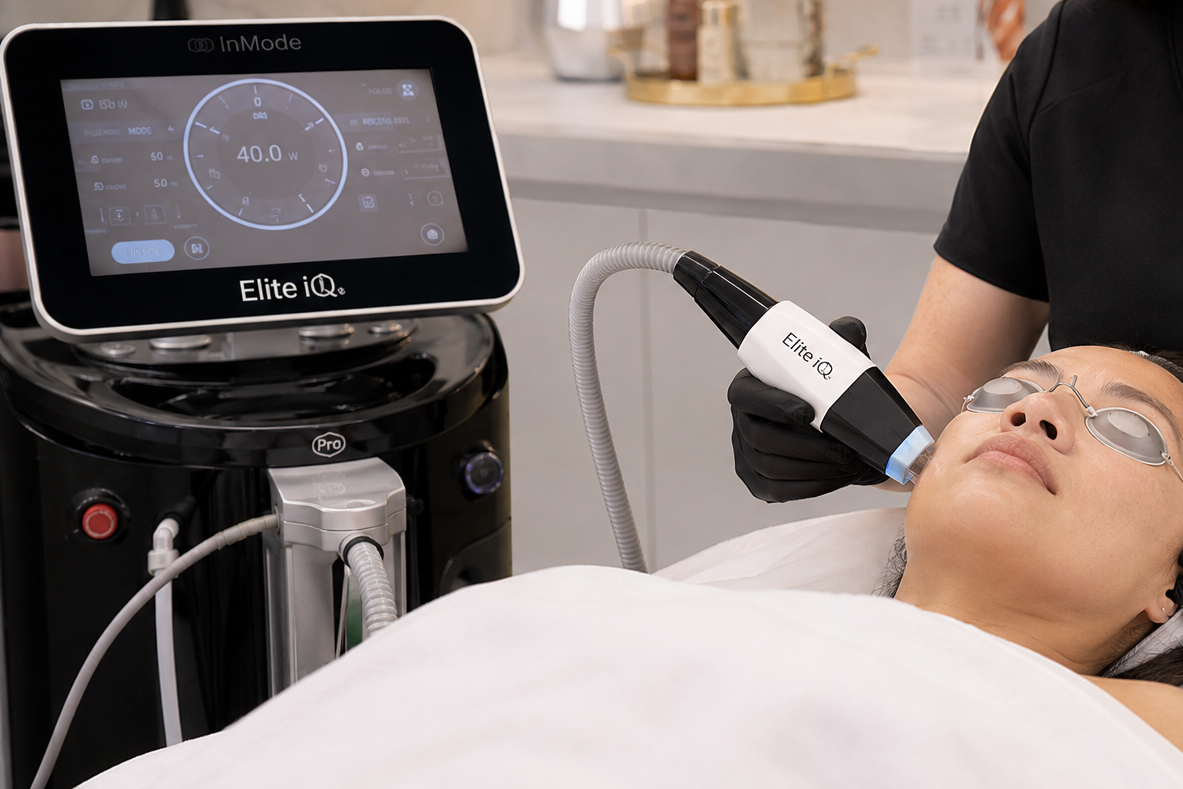 A woman receiving a professional cosmetic treatment with a handheld device connected to a machine labeled Elite iQ, while lying down with protective eye goggles on, in a clinical setting.