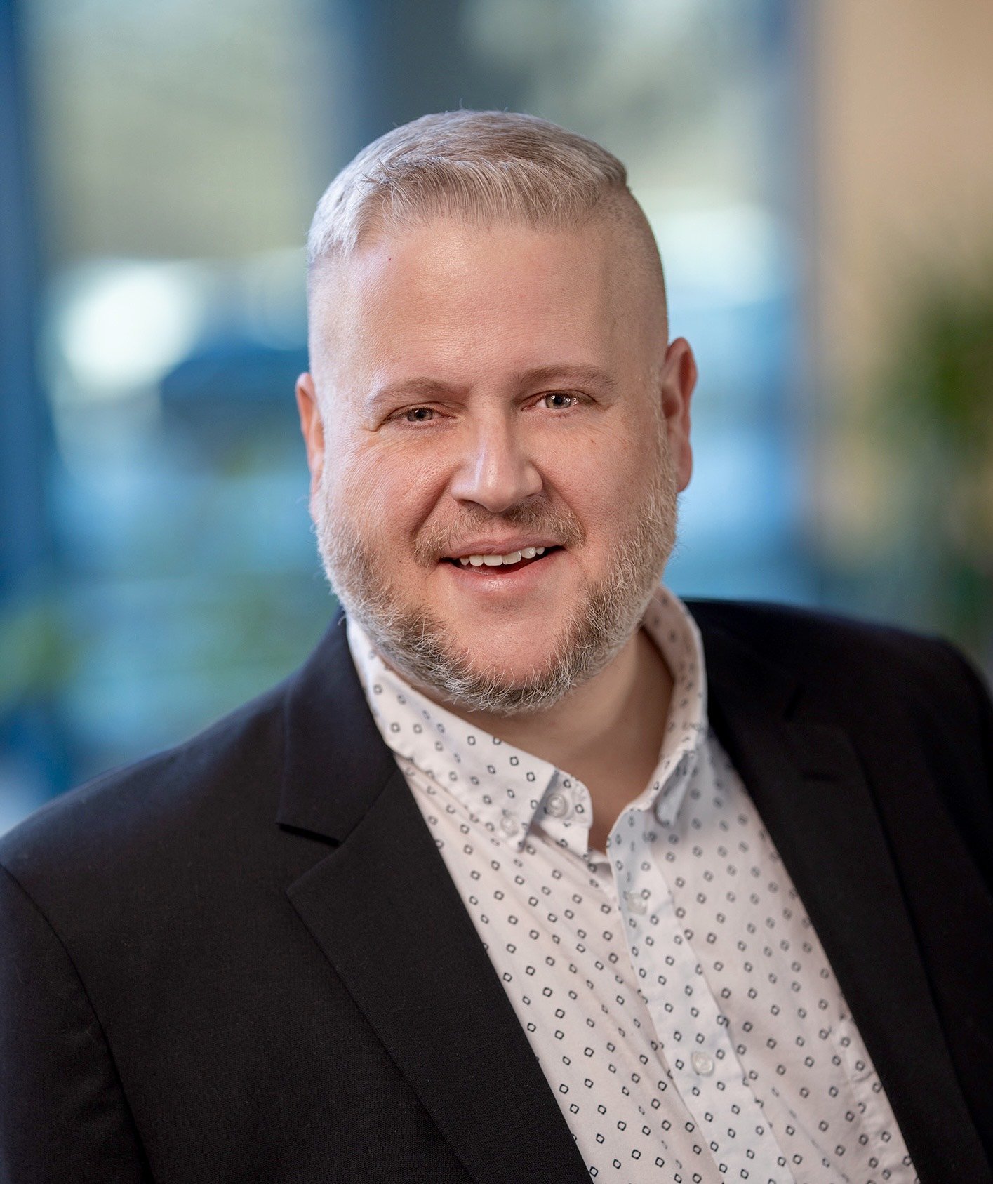 A man with a beard and short hair, wearing a dark suit, light blue dress shirt, and patterned bow tie, smiling and standing against a light-colored wall with a window shadow.