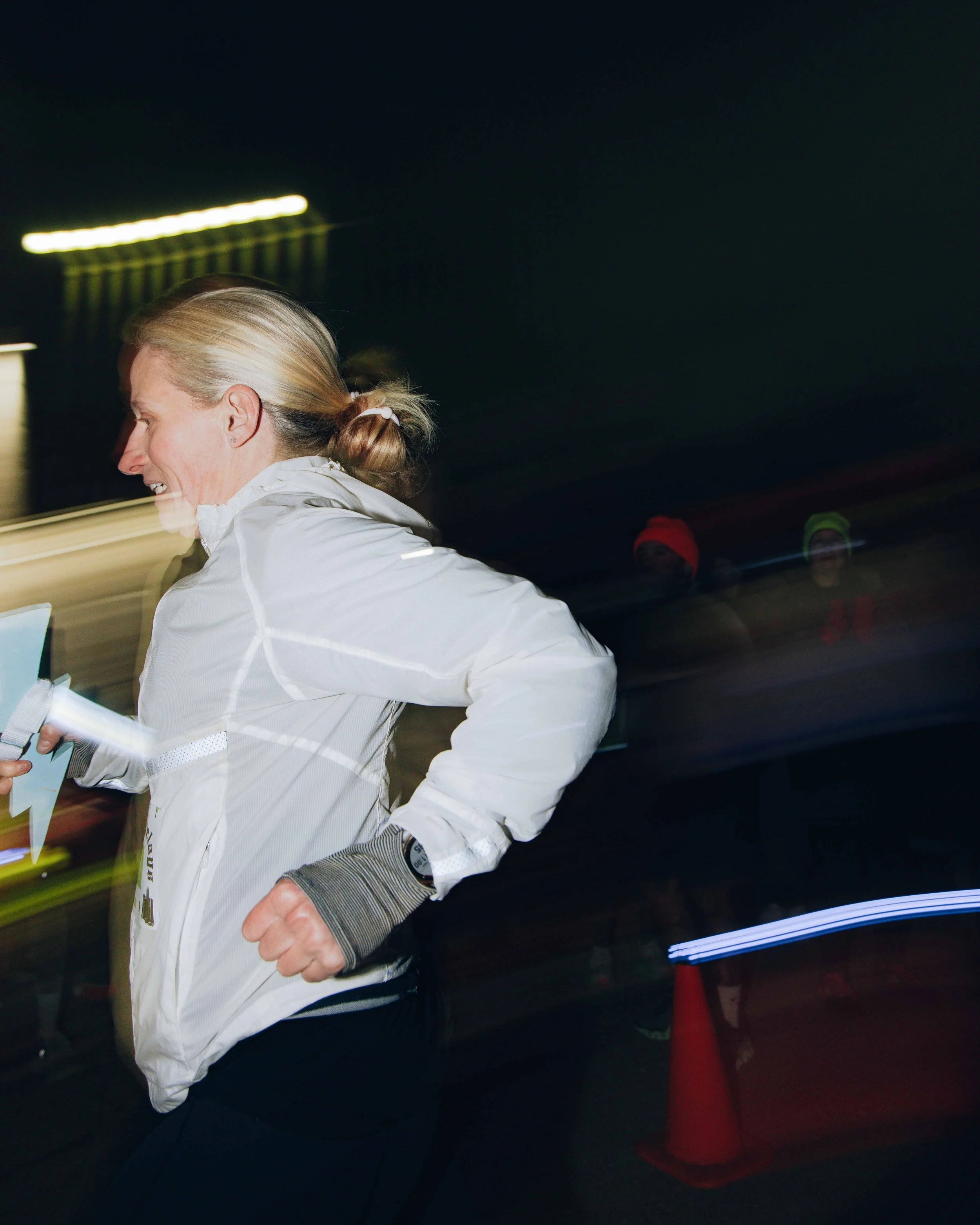 A woman running at night wearing a white jacket, with blurred streaks of light and other runners in the background.