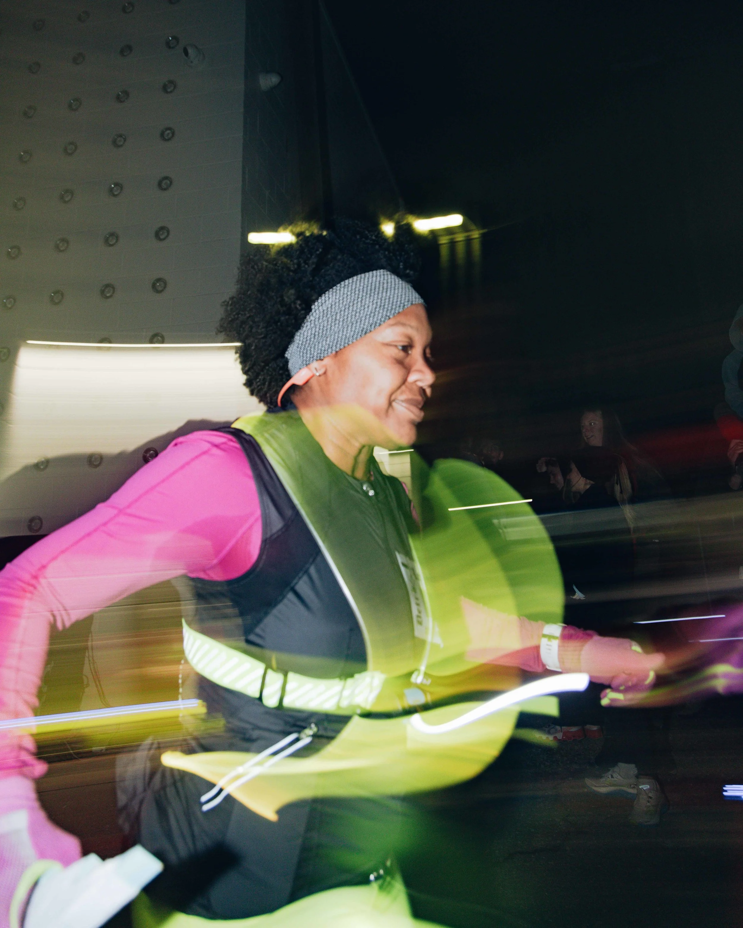 A woman running at night, wearing a pink long sleeve shirt, yellow reflective vest, black shorts, gray headband, and earphones.