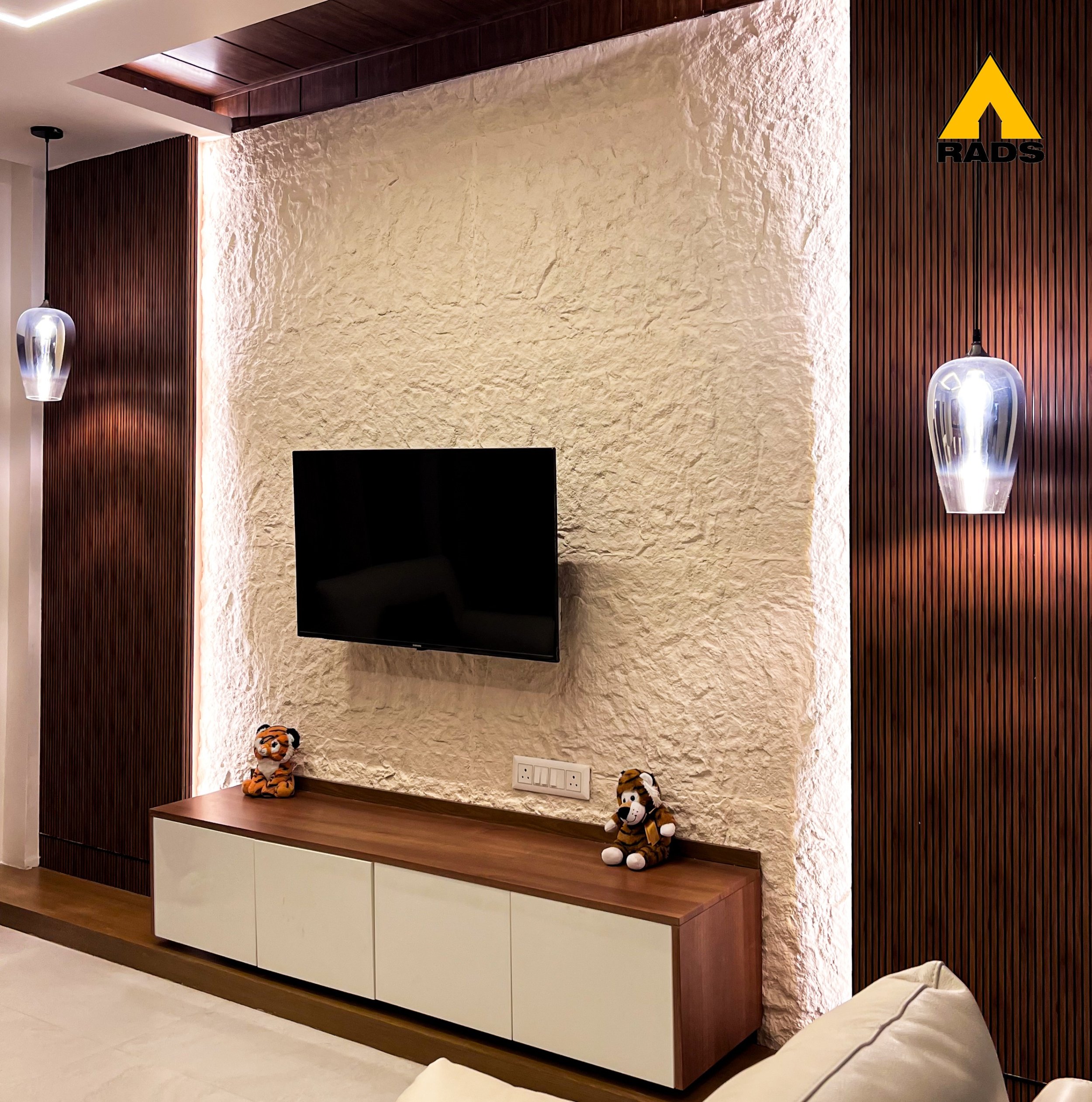 Stylish TV wall unit design with textured cladding and ambient pendant lighting – interior design by RADS in Assam