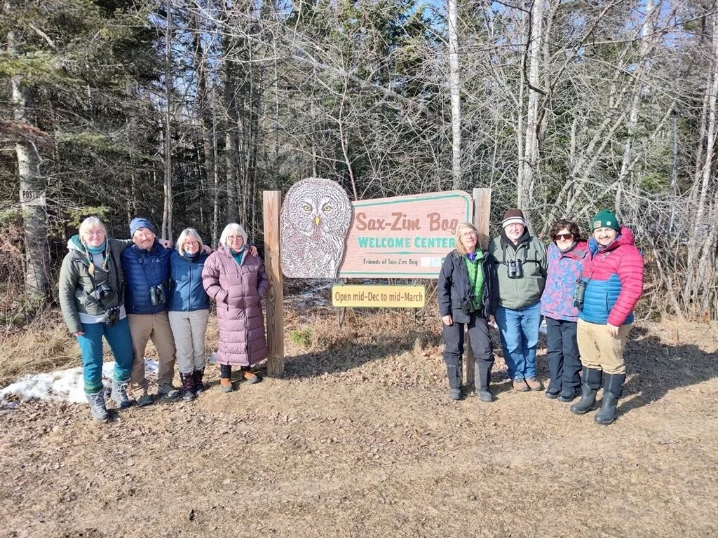 Group of eight people standing in front of a sign at Sax-Zim Bog Welcome Center in a forested area during winter or early spring.