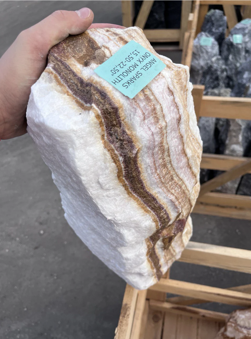 A person holding a large, rectangular block of onyx marble with brown, gold, and white layers, labeled as an onyx marble slab.