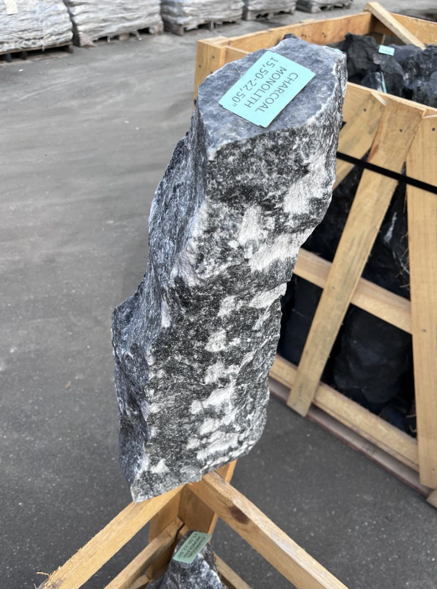 A large, roughly textured gray and white rock with a blue label on top that reads "CHARCOAL MOULD 15-D-22.50." The rock is positioned upright on a wooden stand in a warehouse or outdoor storage area, with other rocks and crates visible in the background.