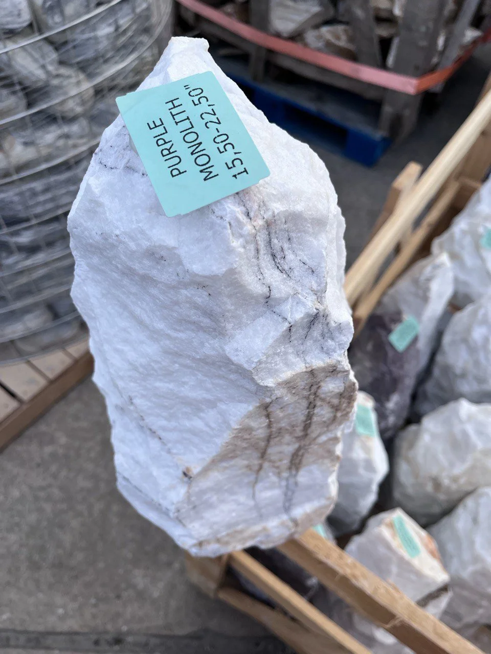 Large chunk of white marble with black veining, labeled 'Purple Monnolith' and a serial number, displayed outdoors in a store or warehouse with other stones and wood visible in the background.