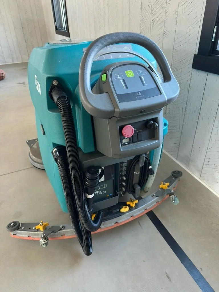 A rideable floor cleaning machine with a handle, control panel, and cleaning brushes, positioned on a concrete floor near a wall and window.