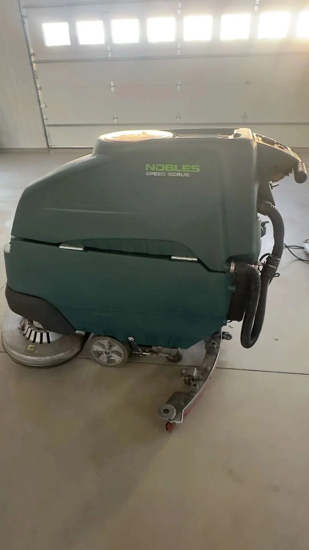 A Nobles Speed Scrub floor cleaning machine in a garage with a concrete floor and a white garage door in the background.