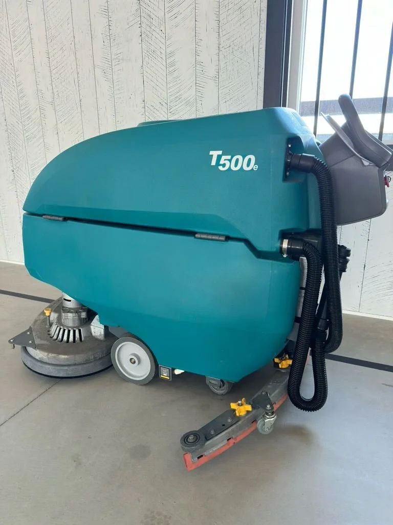 Commercial floor cleaning machine, T500e model, with large blue body, black hoses, and cleaning brushes on a concrete floor near a window.