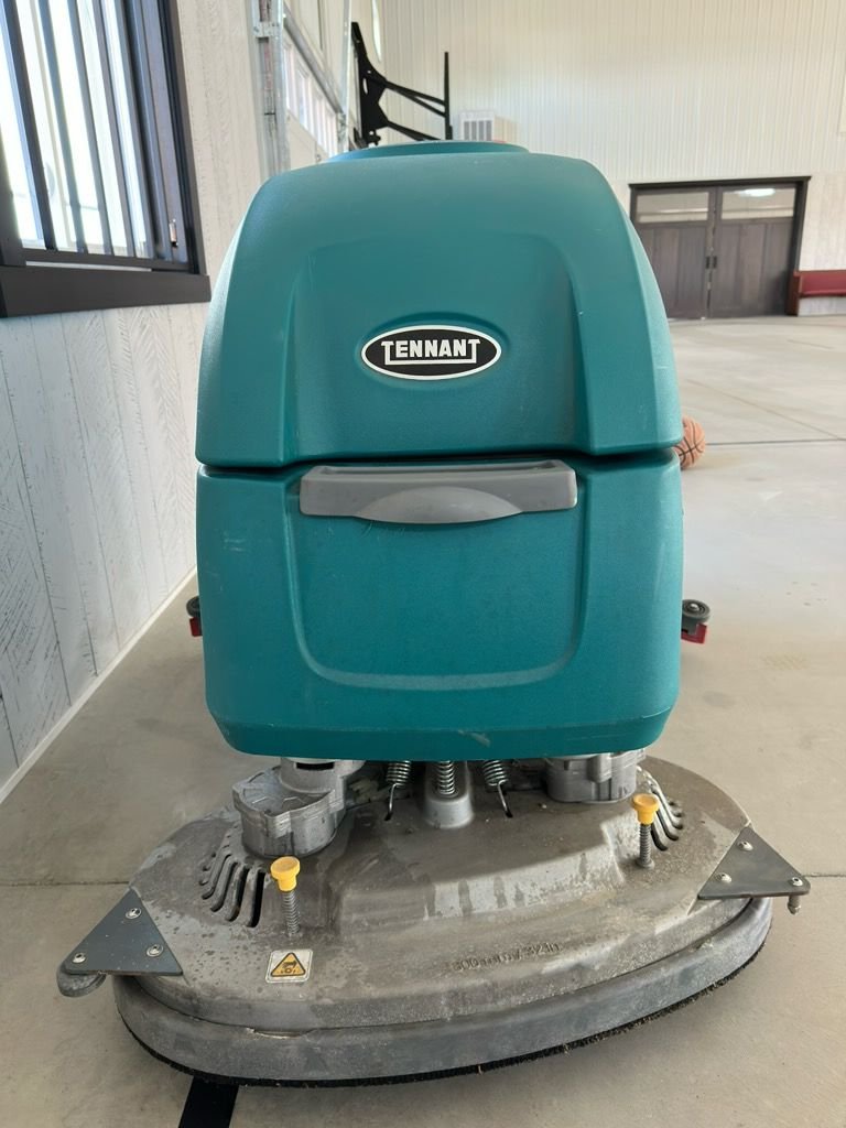 A Tennant brand floor cleaning machine inside a building, with a teal body and a gray base, placed on a concrete floor near a wall and window.