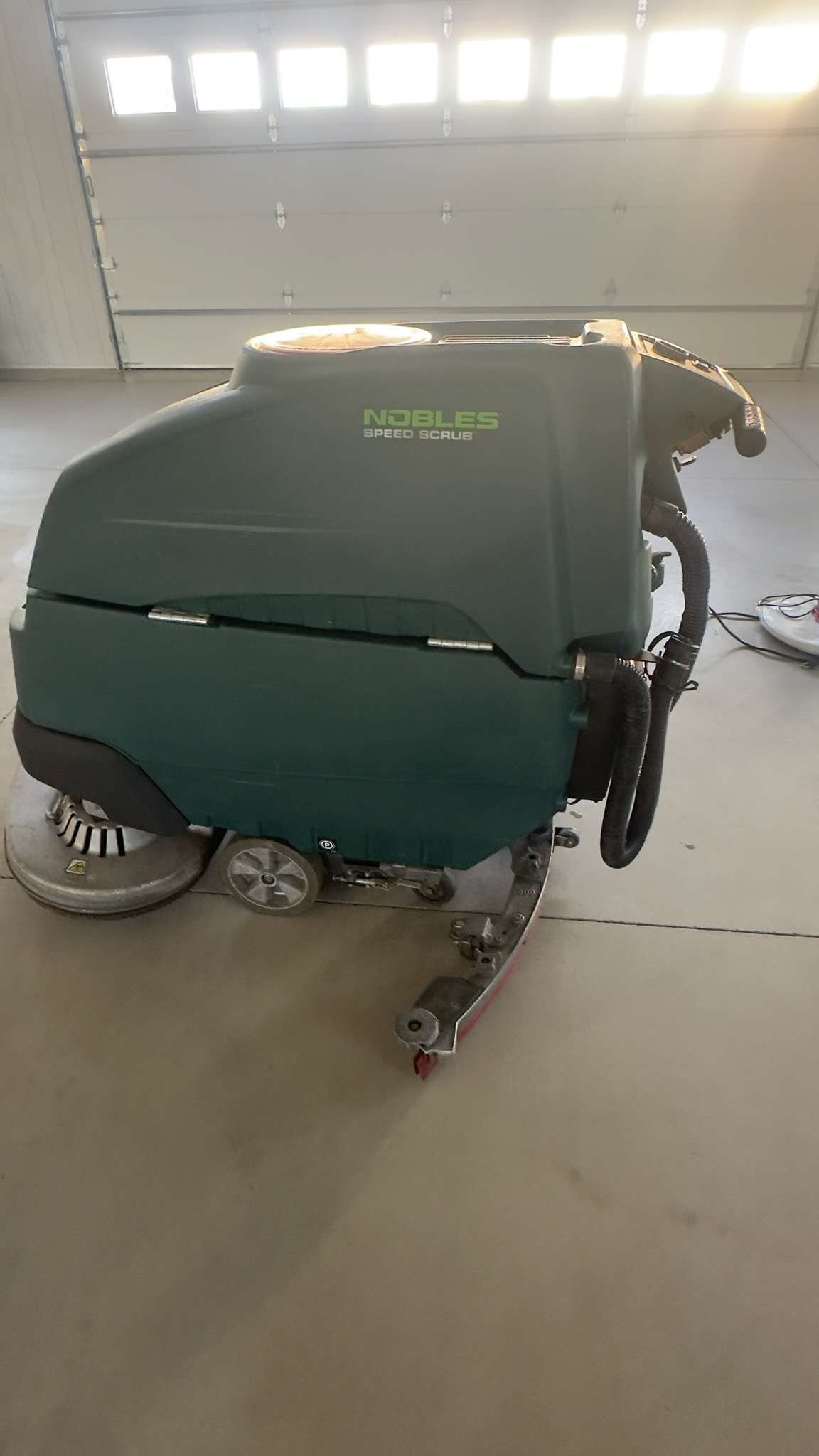 A green Nobles Speed Scrub floor cleaning machine on a concrete garage floor with a closed garage door in the background.