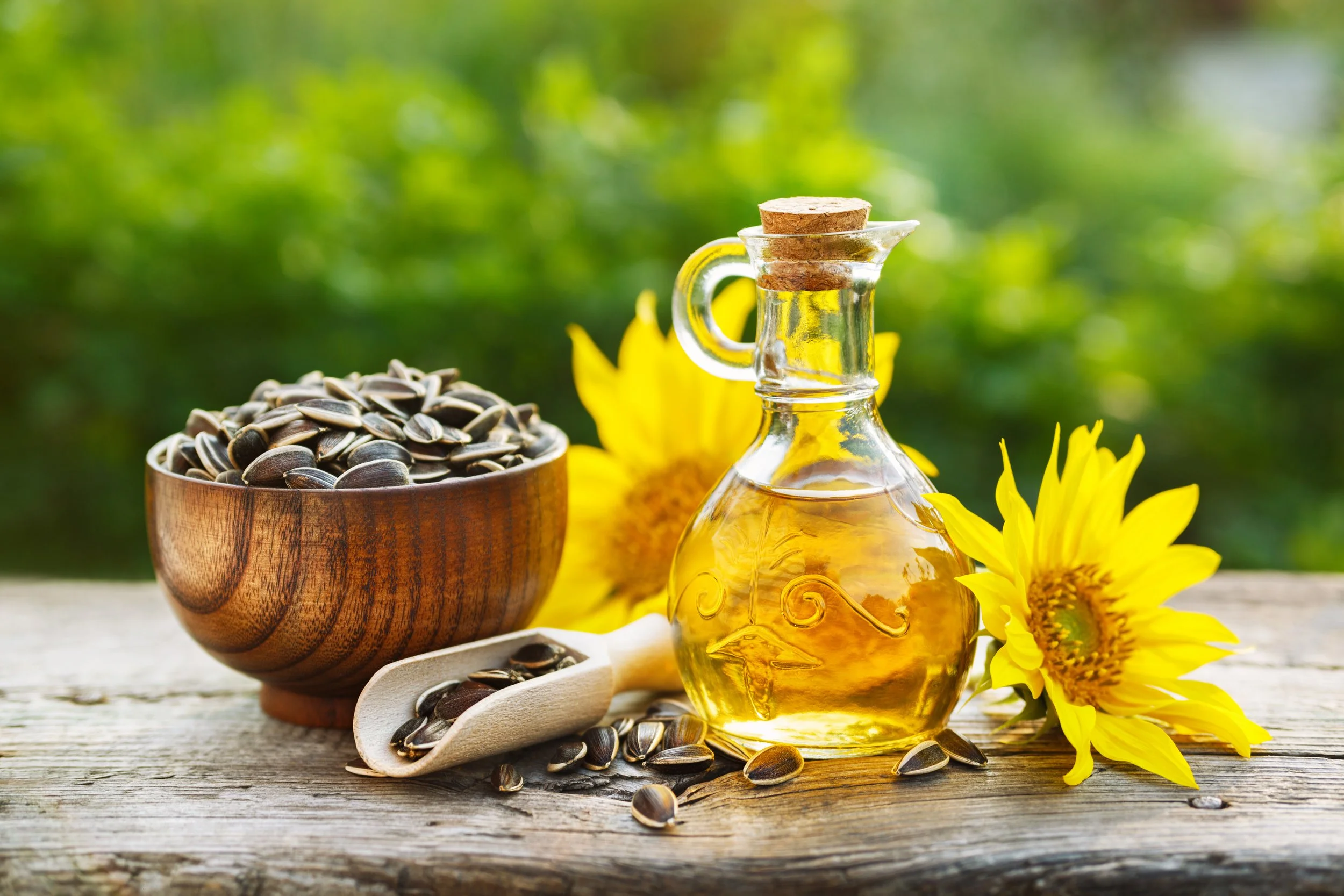 Sunflowers, a glass bottle of sunflower oil with a cork, a wooden bowl of sunflower seeds, and a scoop of sunflower seeds on a rustic wooden surface with a blurred green background.