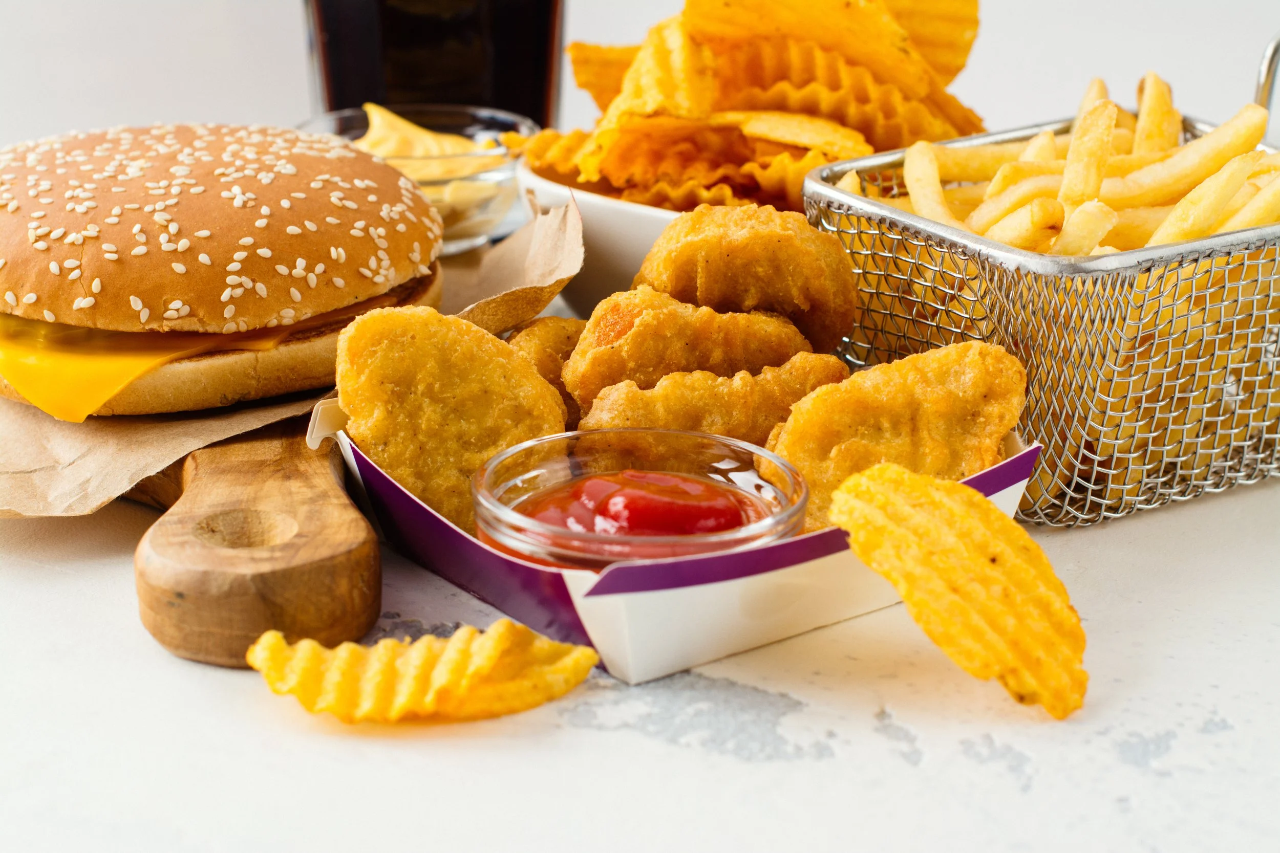 A fast food meal featuring a cheeseburger with pickles and cheese, a basket of French fries, a container of chicken nuggets with a side of ketchup, and a bowl of potato chips.