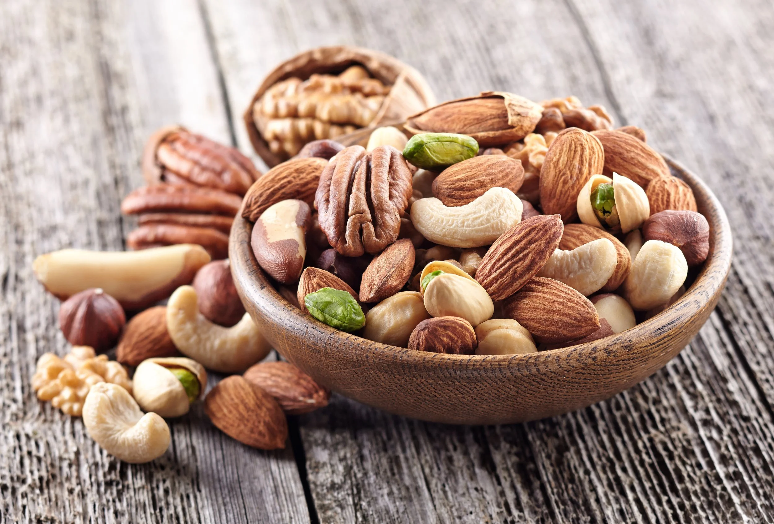 A wooden bowl filled with mixed nuts, including almonds, cashews, pecans, hazelnuts, and pistachios, on a rustic wooden surface.