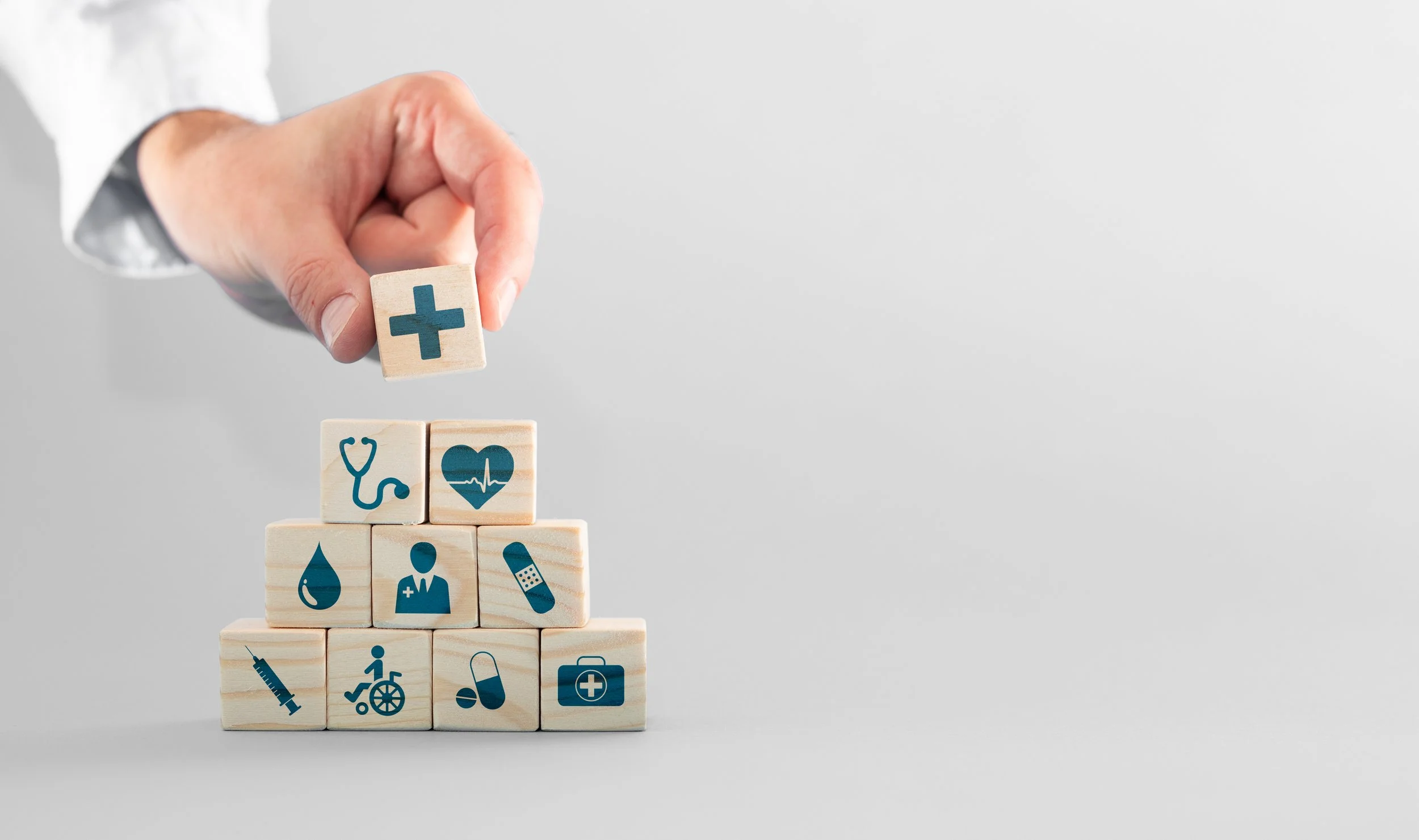 A hand placing a wooden block with a medical cross on a pyramid of blocks with healthcare icons.