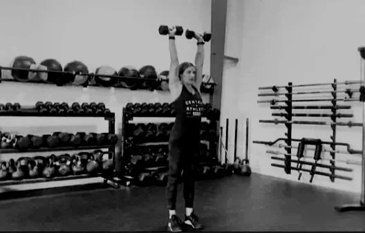Woman lifting two dumbbells overhead in a gym.