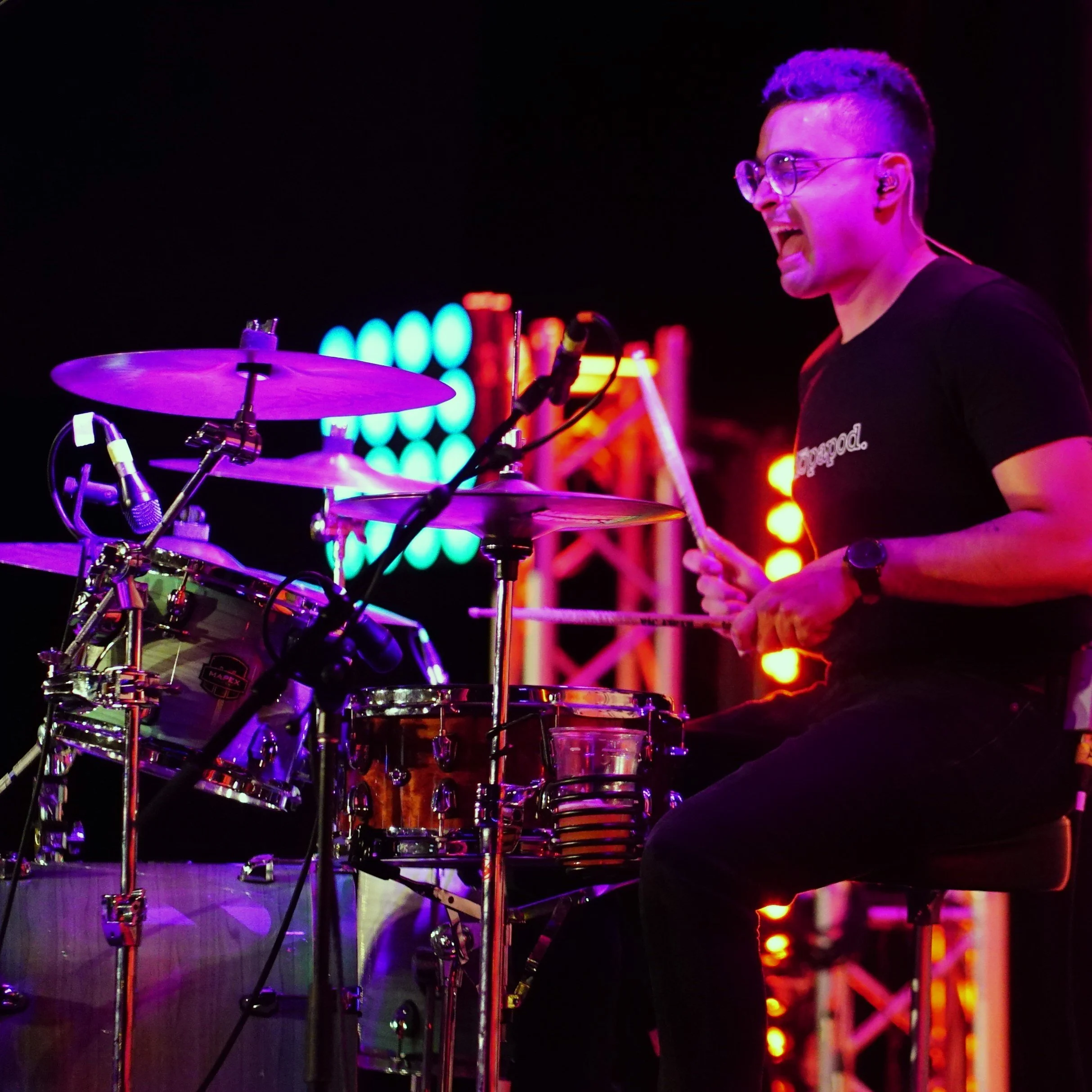 A woman singing into a microphone on stage, wearing a strapless dress and tall black boots, with a guitar and drums in the background, and an American flag with stars in the background.