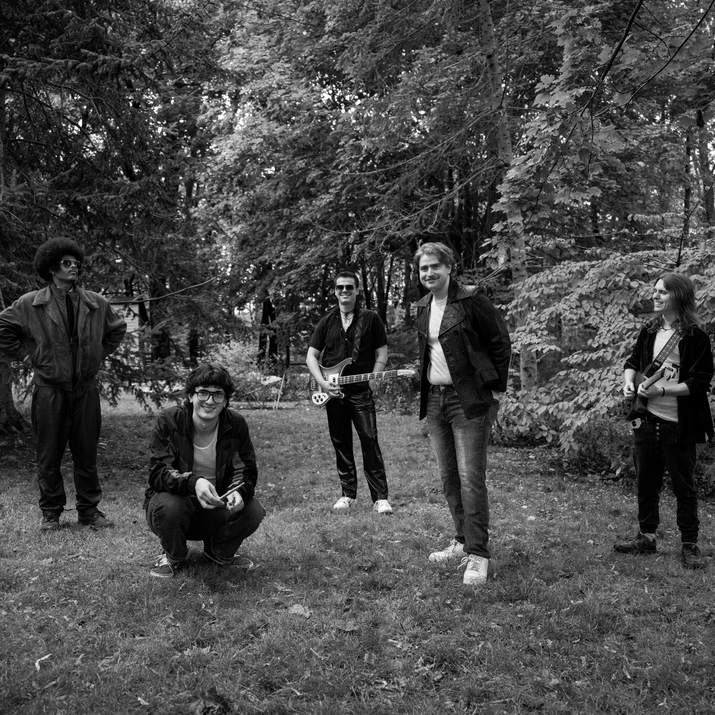 A black-and-white photo of a group of five people outdoors in a wooded area, some holding guitars and others posing casually, smiling and dressed in casual clothing.