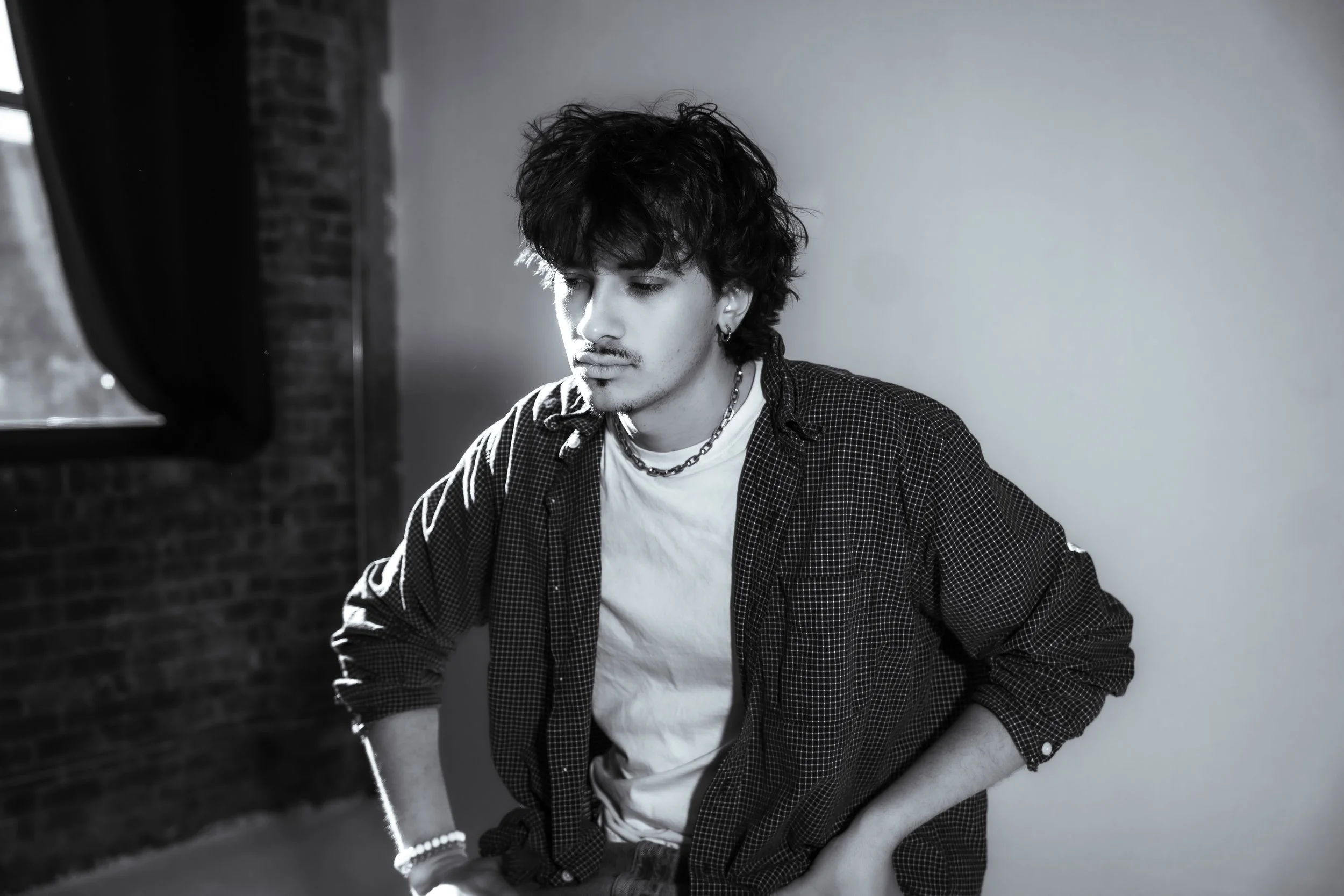 A young man with curly dark hair and earrings, wearing a checkered shirt over a white T-shirt, sitting indoors near a window with brick wall, looking down with a contemplative expression.