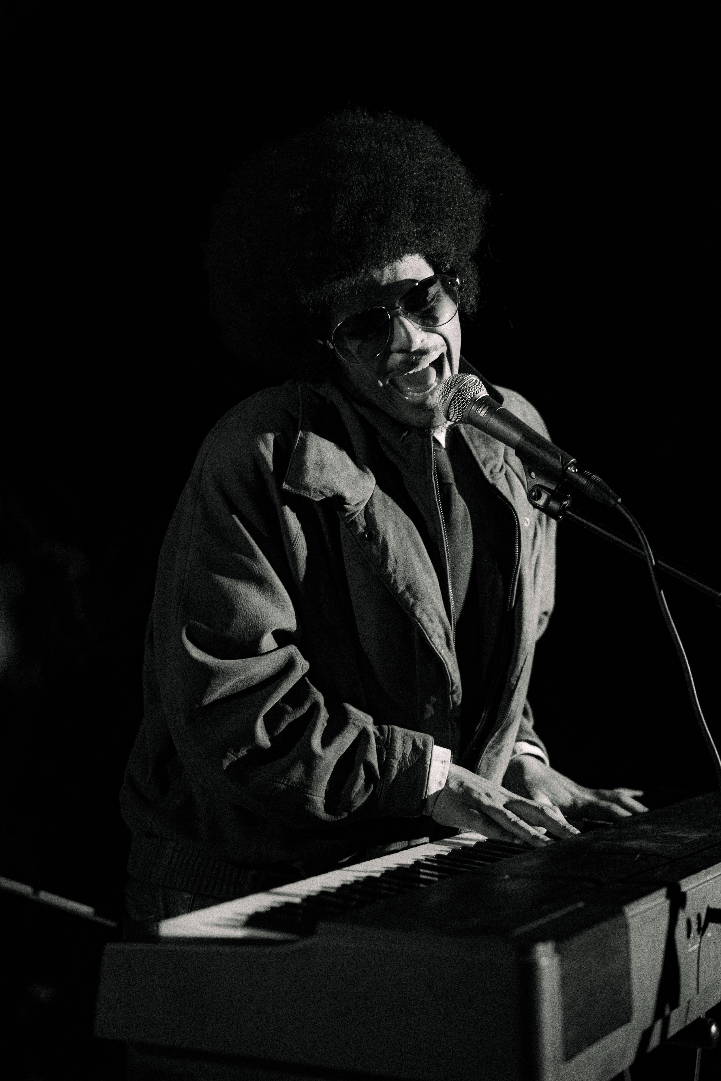 A black and white photo of a musician singing and playing keyboard on stage, wearing sunglasses and a jacket.