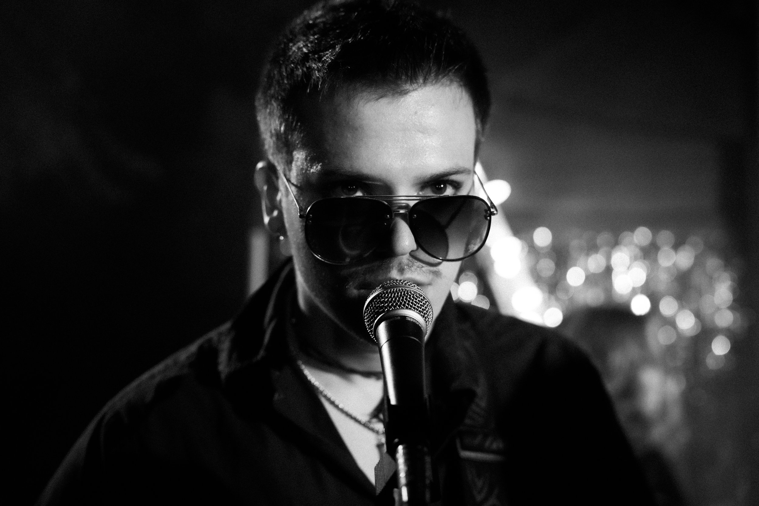A person with short hair wearing sunglasses, speaking into a microphone in a dark setting with blurred lights in the background.