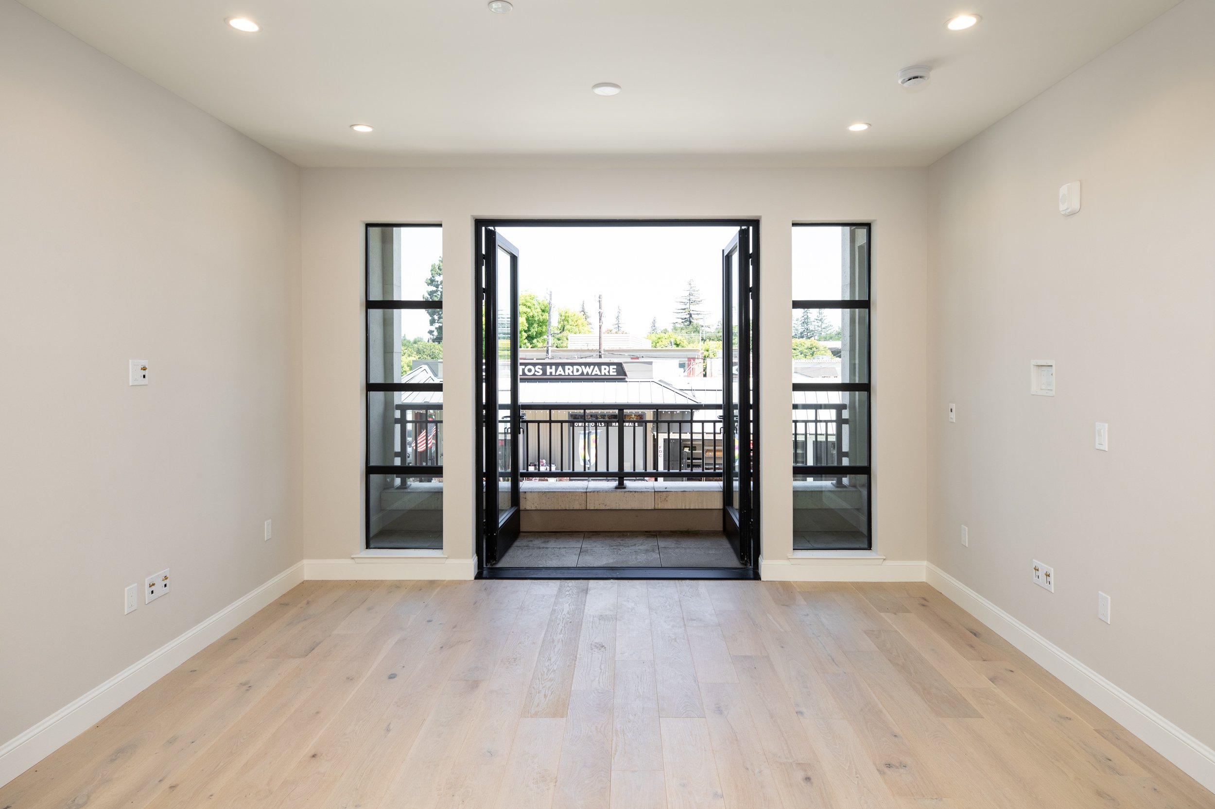 Empty room with light wood flooring, white walls, and a black-framed open balcony door leading to a small outdoor space with a railing. Outside view includes a commercial building with a sign that reads 'HARDWARE' and some trees in the background.