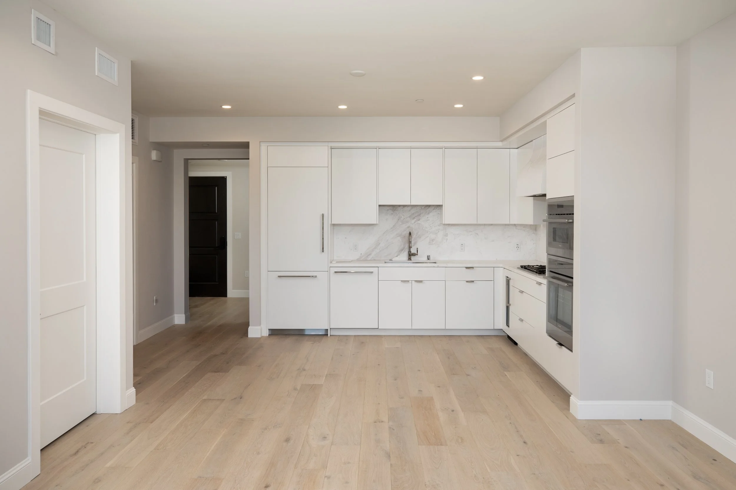 Modern, minimalist white kitchen with light wood flooring, marble backsplash, and built-in appliances.