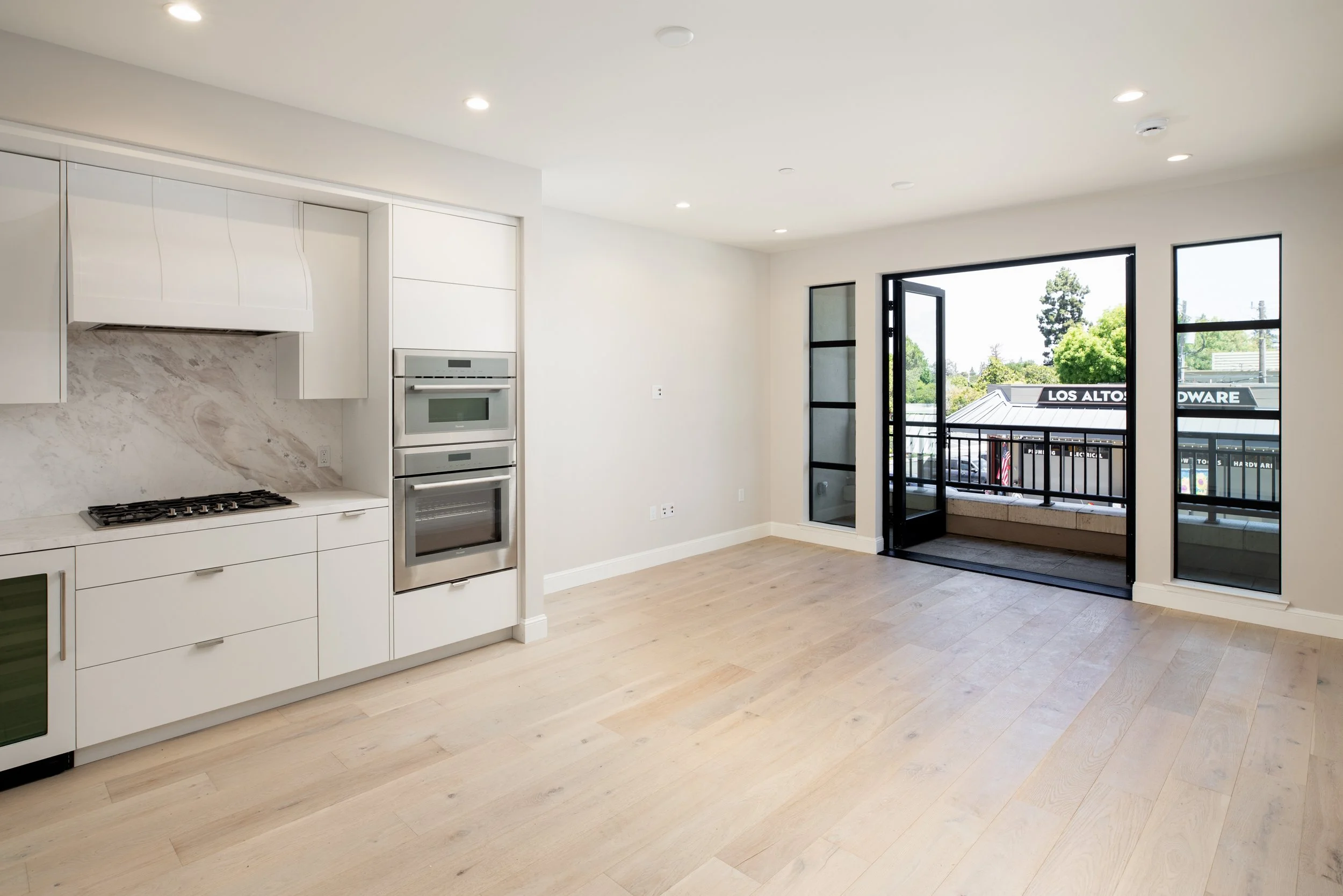 Empty modern apartment living area with white cabinetry kitchen, light wood flooring, and large glass door leading to balcony with neighborhood view.