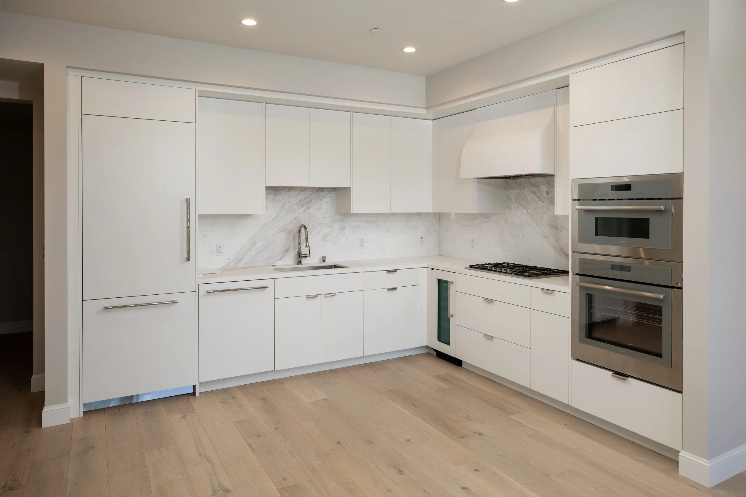 Modern white kitchen with stainless steel oven and microwave, marble backsplash, and light wood flooring.
