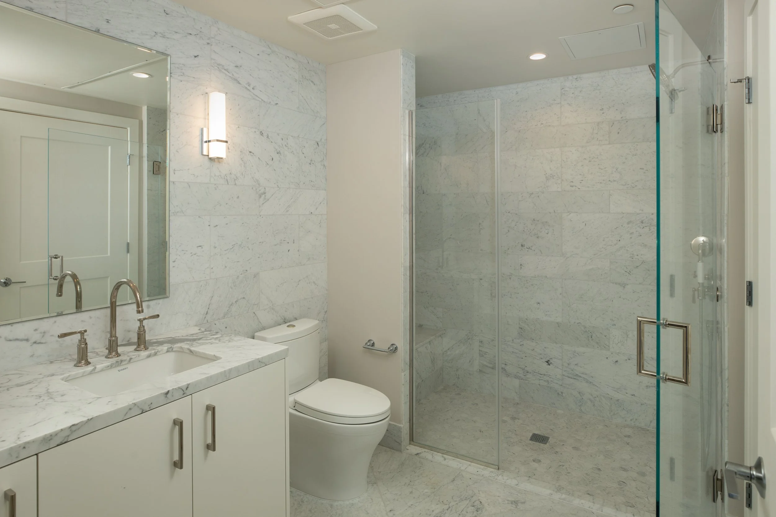 Modern bathroom with white marble walls and floor, a white vanity with a marble countertop and chrome fixtures, a wall-mounted light, a toilet, and a glass-enclosed shower with marble walls.