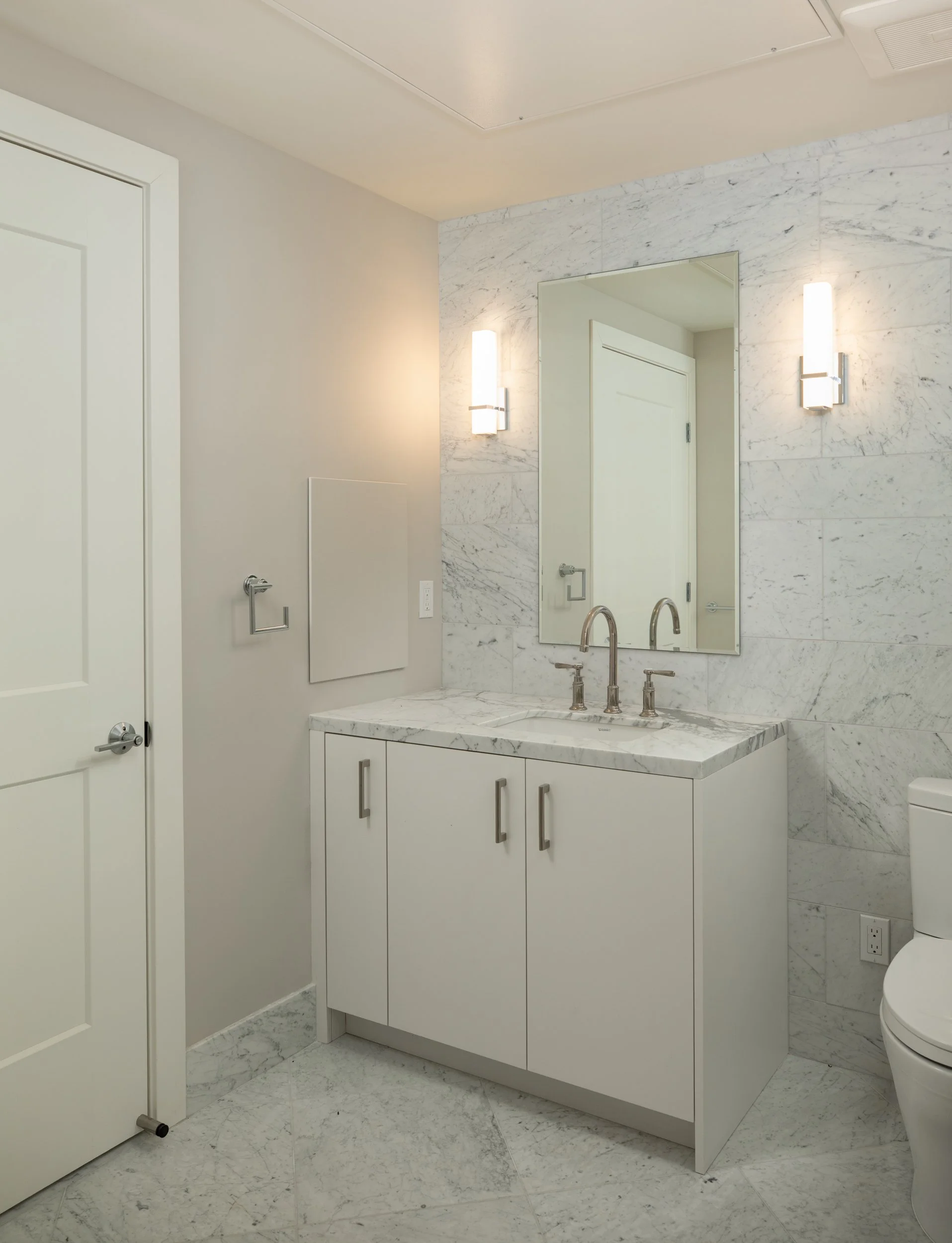 Modern bathroom with white vanity, marble countertop, large mirror, wall-mounted lights, and toilet.