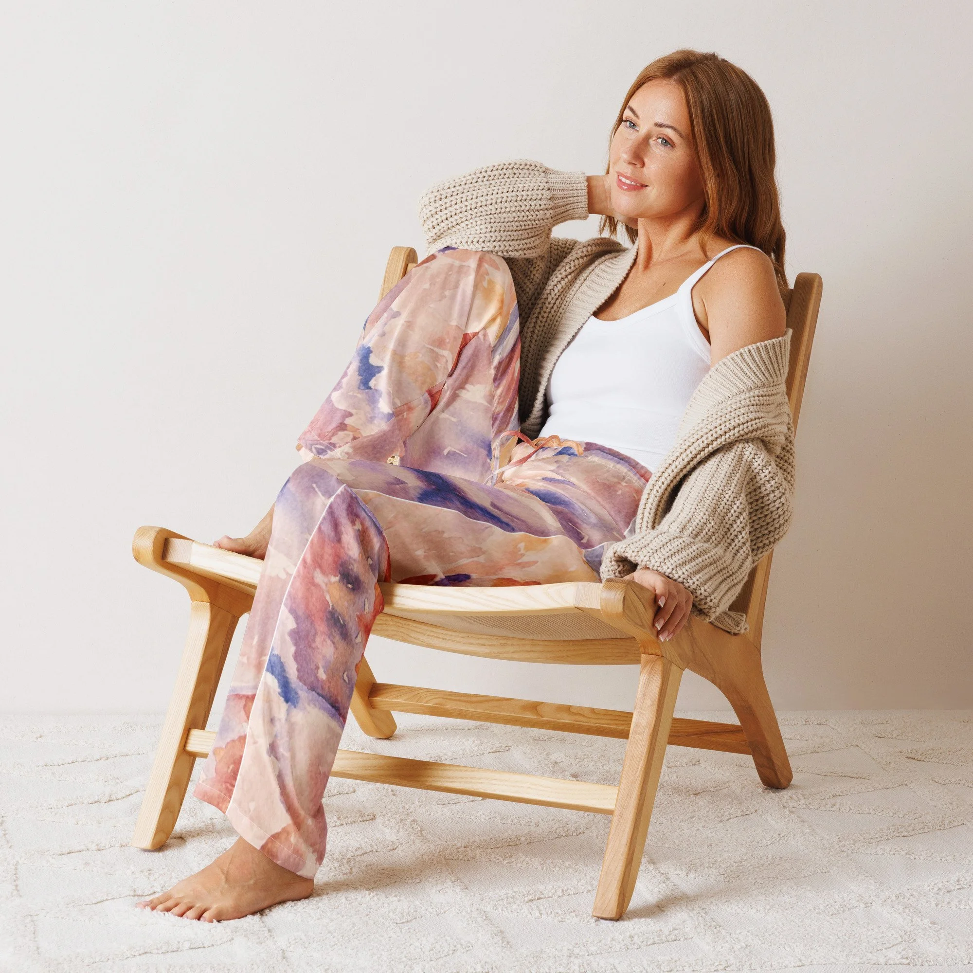 A woman with red hair sitting casually on a wooden chair, wearing colorful patterned pants, a white tank top, and a beige knitted cardigan, with a relaxed expression.