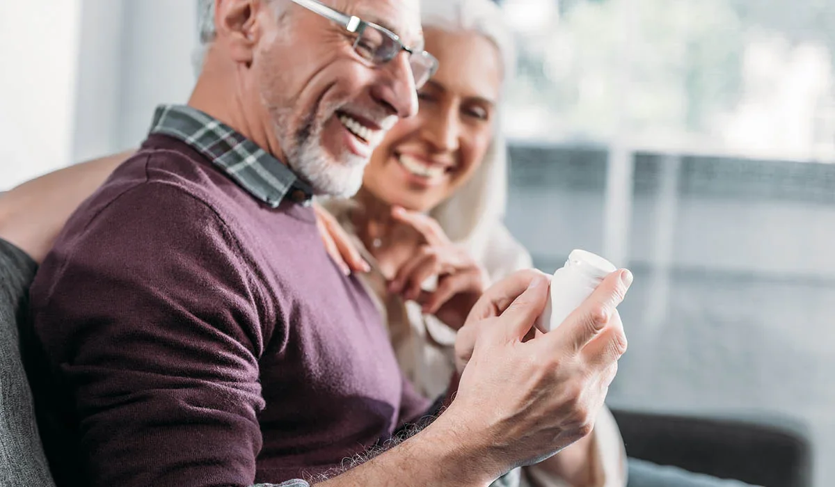An elderly man and woman sitting together, smiling, with the man holding a small white container in his hand.