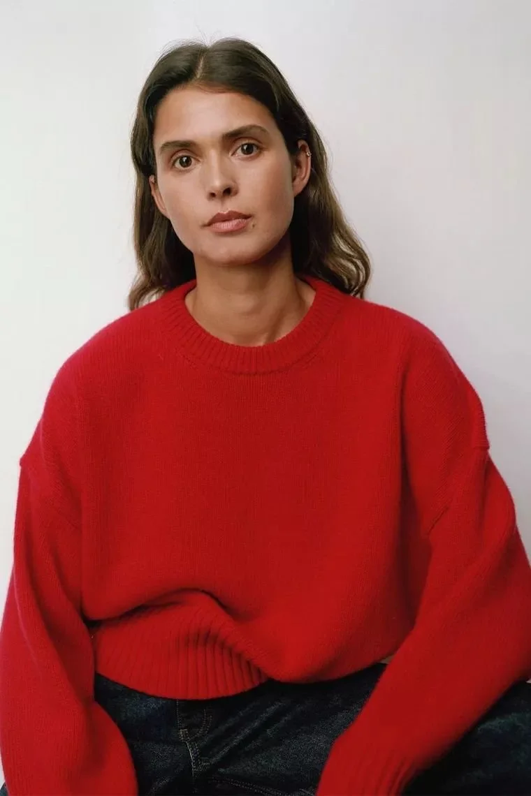 Femme aux cheveux bruns, portant un pull rouge et assise contre un mur blanc.