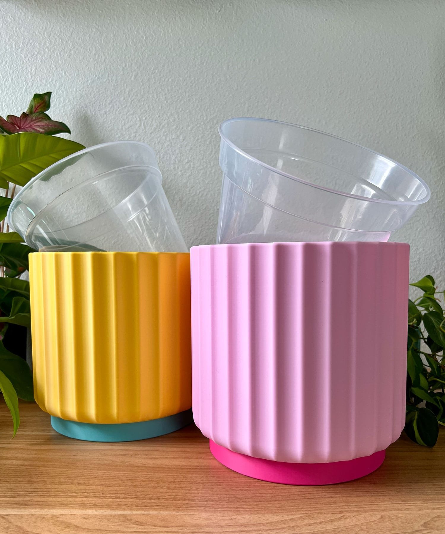 2 large 8 inch colorful planters sitting on a wood table against a cream colored wall. planters are sunny yellow and light pink with clear liner pots sticking out of the top
