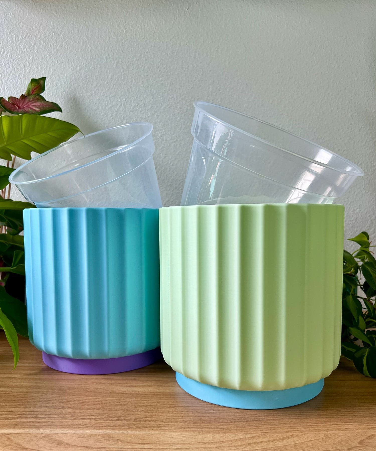 2 large 8 inch planters sitting on a wood table against a cream colored wall. planters are light blue and green with clear liner pots sticking out of the top at different angles.
