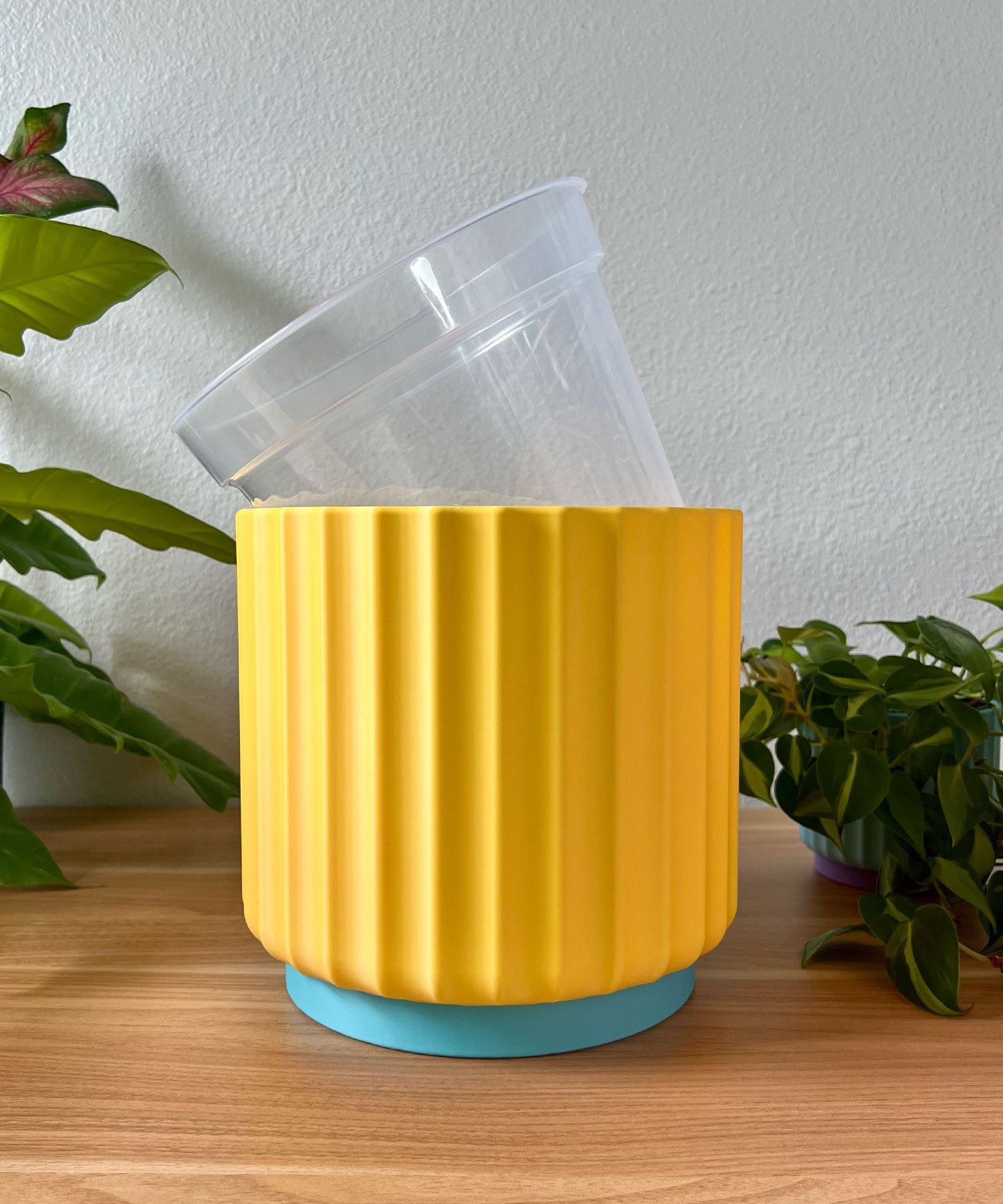 large 8 inch sunny yellow planter with a clear liner pot sitting on a wood table against a cream colored wall with green plants in the background