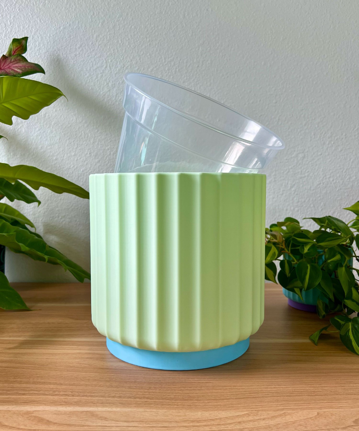 large 8 inch light green planter with a clear liner pot sitting on a wood table against a cream colored wall with green plants in the background