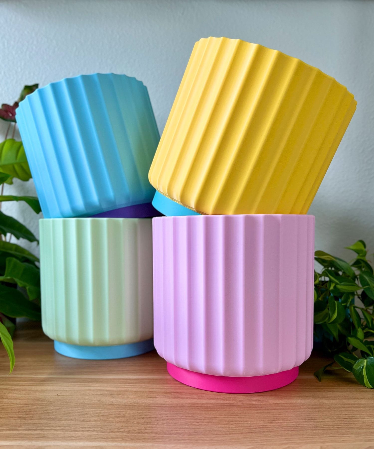 4 large 8 inch planters sitting in 2 stacked rows on a wood table, against a white wall. sunny yellow, light pink, light blue, and light green with green plants in the background