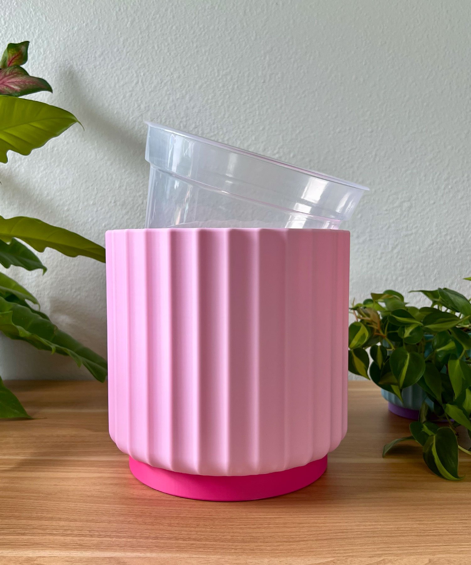 large 8 inch light pink planter with a clear liner pot sitting on a wood table against a cream colored wall with green plants in the background