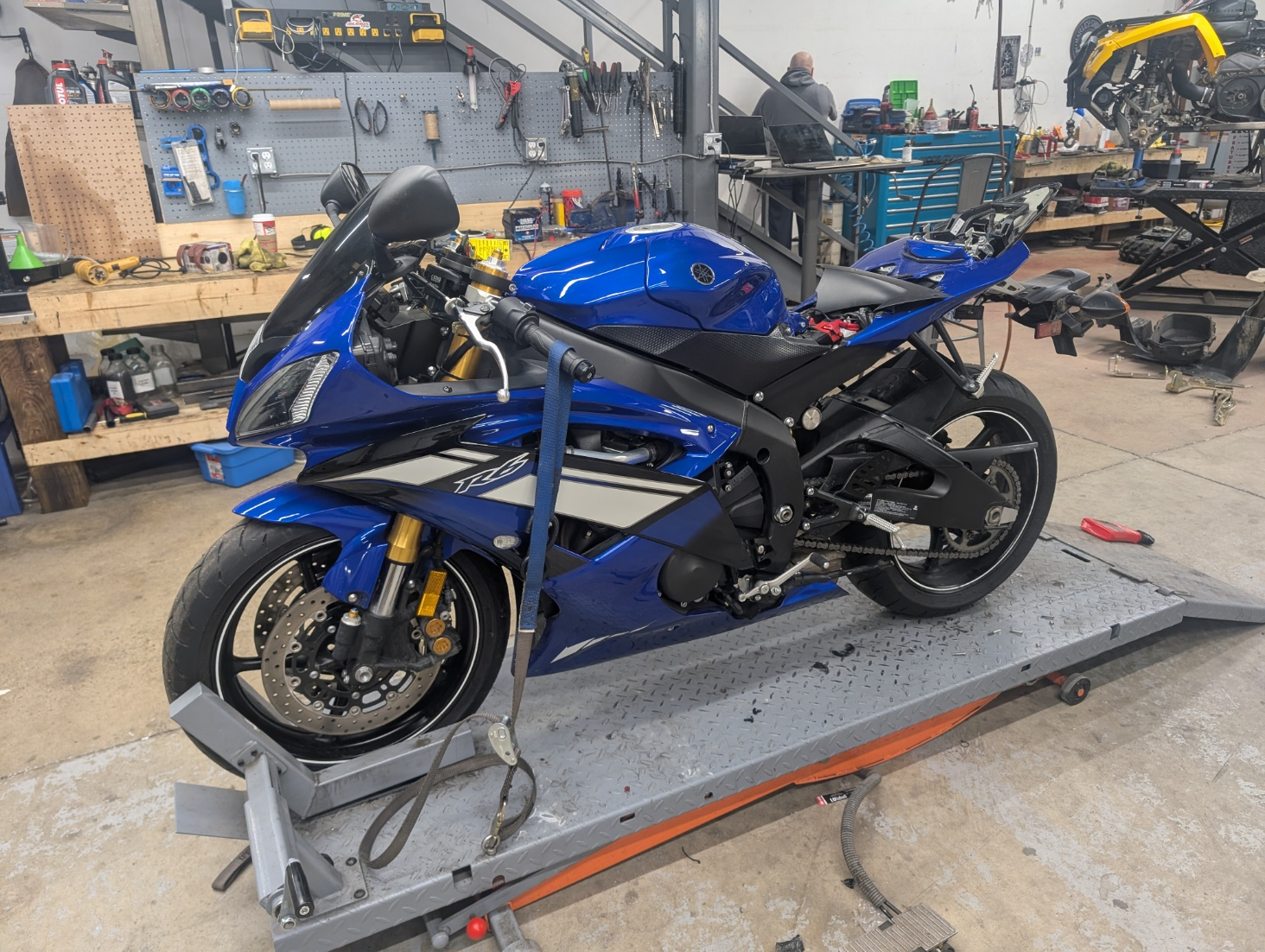 A blue Yamaha YZF-R6 motorcycle on a work stand inside a workshop, surrounded by various tools and motorcycle parts.
