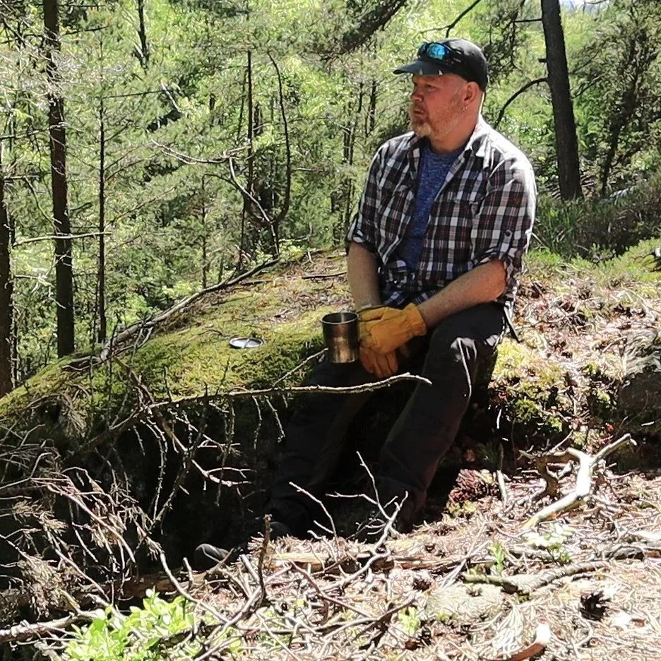 A man sitting on a moss-covered log in a forest, wearing a plaid shirt, black pants, yellow gloves, and a black cap with sunglasses, holding a metal mug, surrounded by trees and fallen branches.