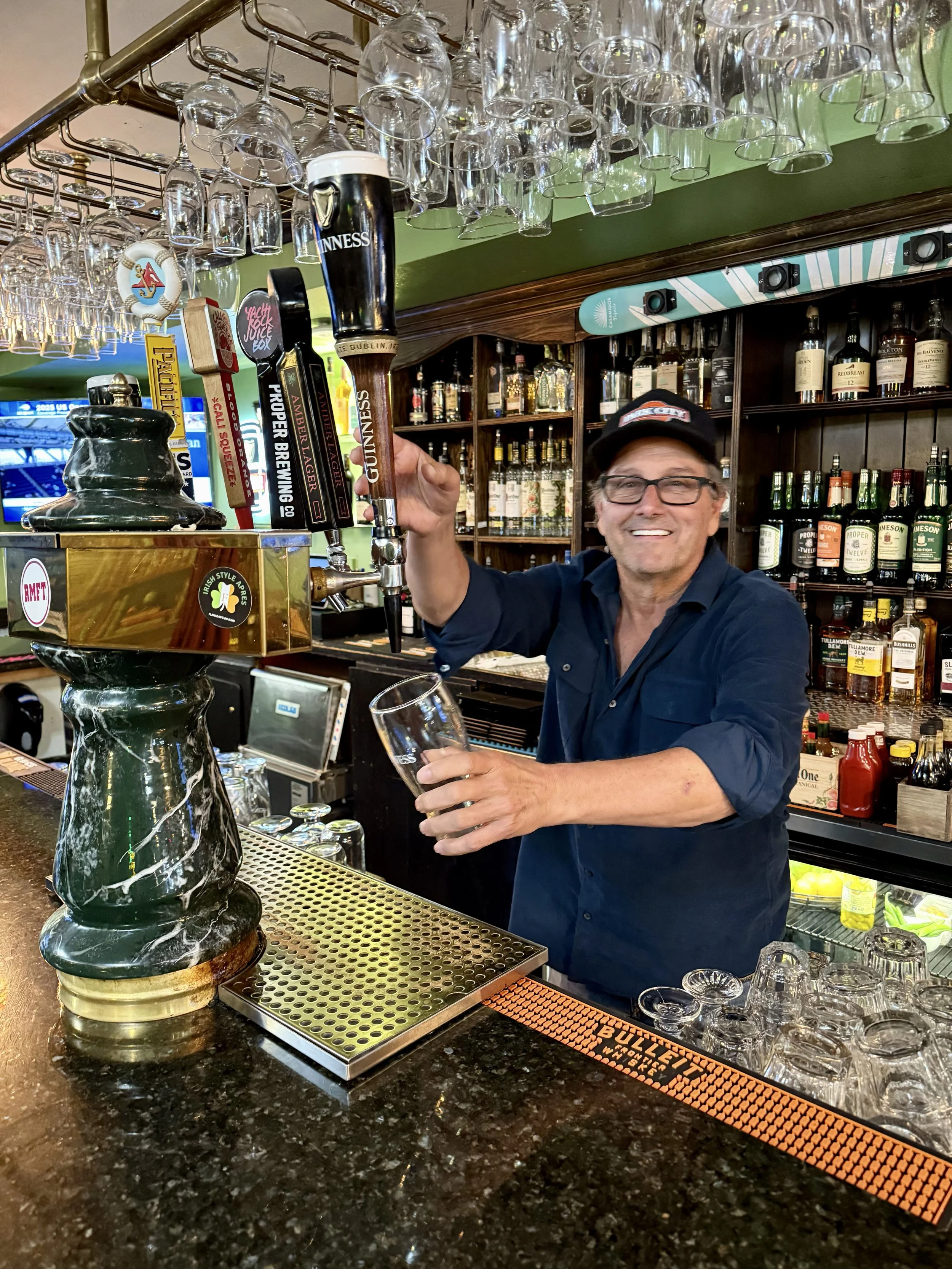 A smiling bartender in a navy shirt and glasses pours a draft beer from a tap into a glass at a bar with shelves of liquor bottles in the background.