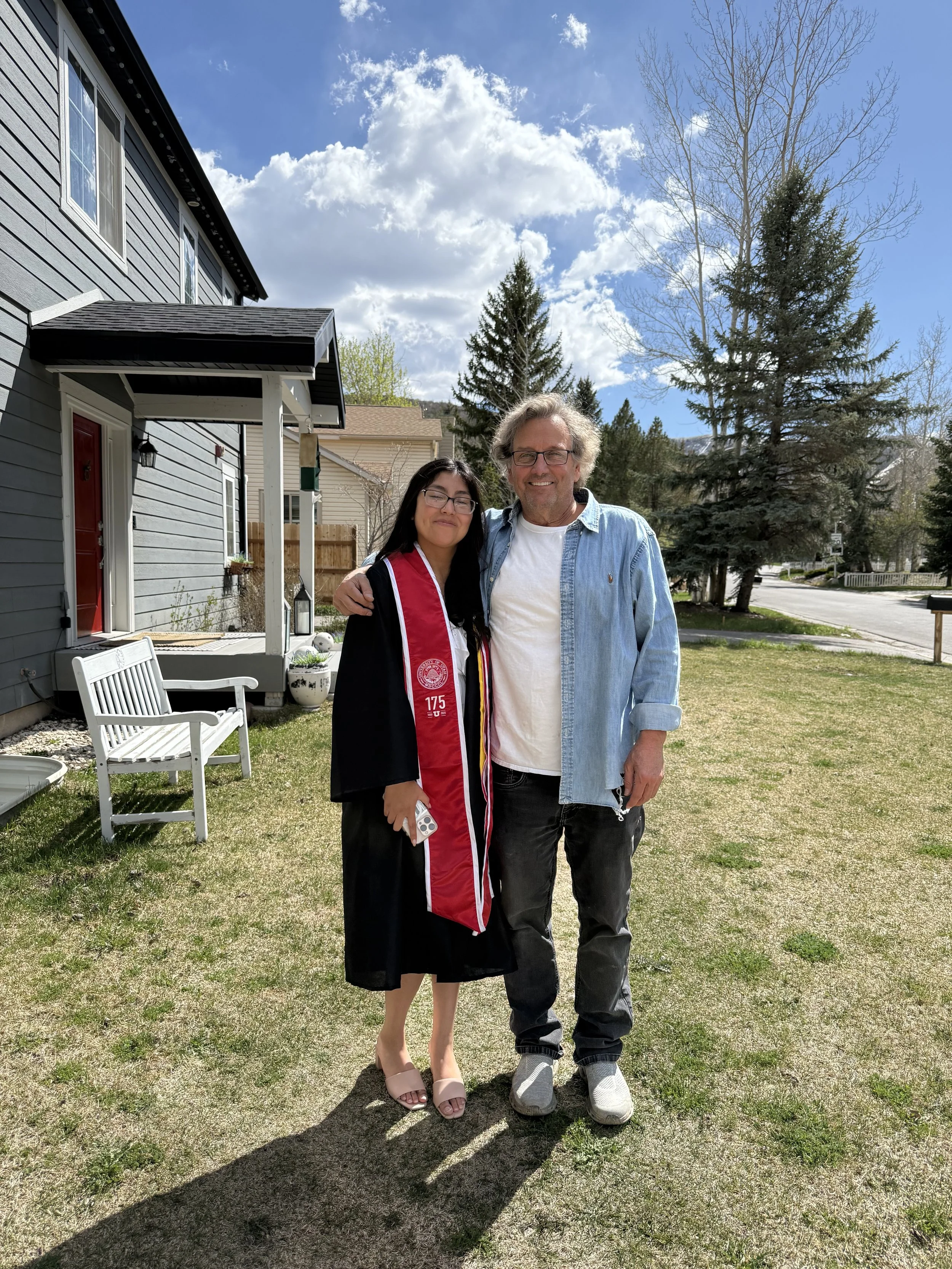 A young woman in a graduation cap and gown with a red stole stands next to an older man in casual clothing in a front yard on a sunny day with clouds. They are smiling and pose with arms around each other.