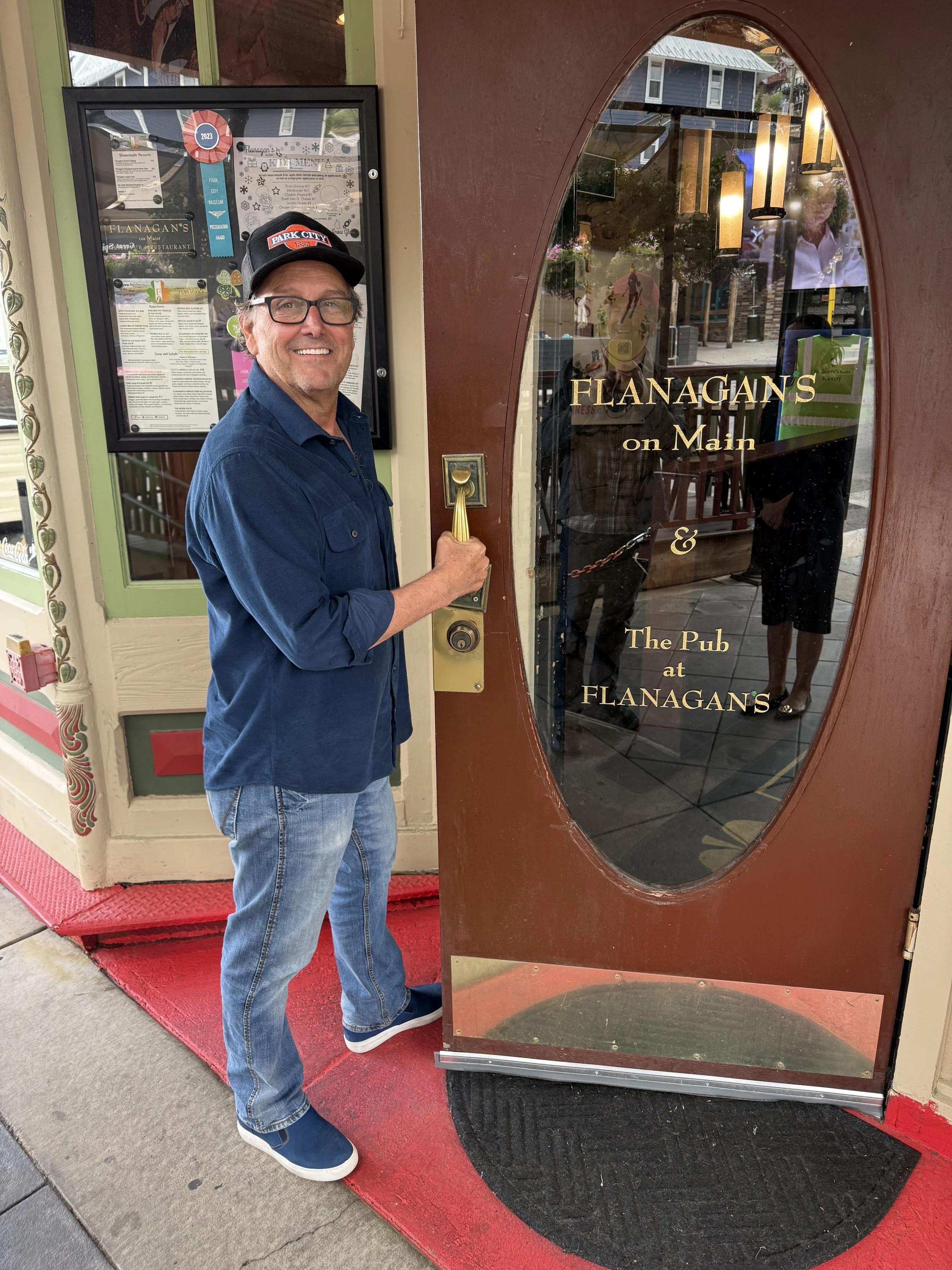 A man smiling and holding the door handle of a restaurant called Flanagan's on Main. The restaurant has a glass door with gold lettering, and the man is wearing a blue shirt, jeans, a black cap, and glasses. Reflection of people and the street can be seen in the glass door.