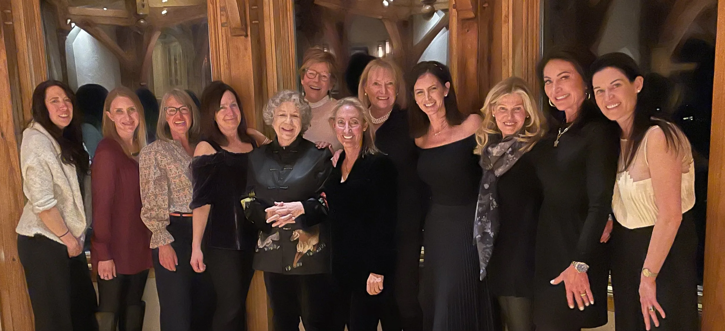 A group of women smiling together indoors, standing closely in front of wooden walls.