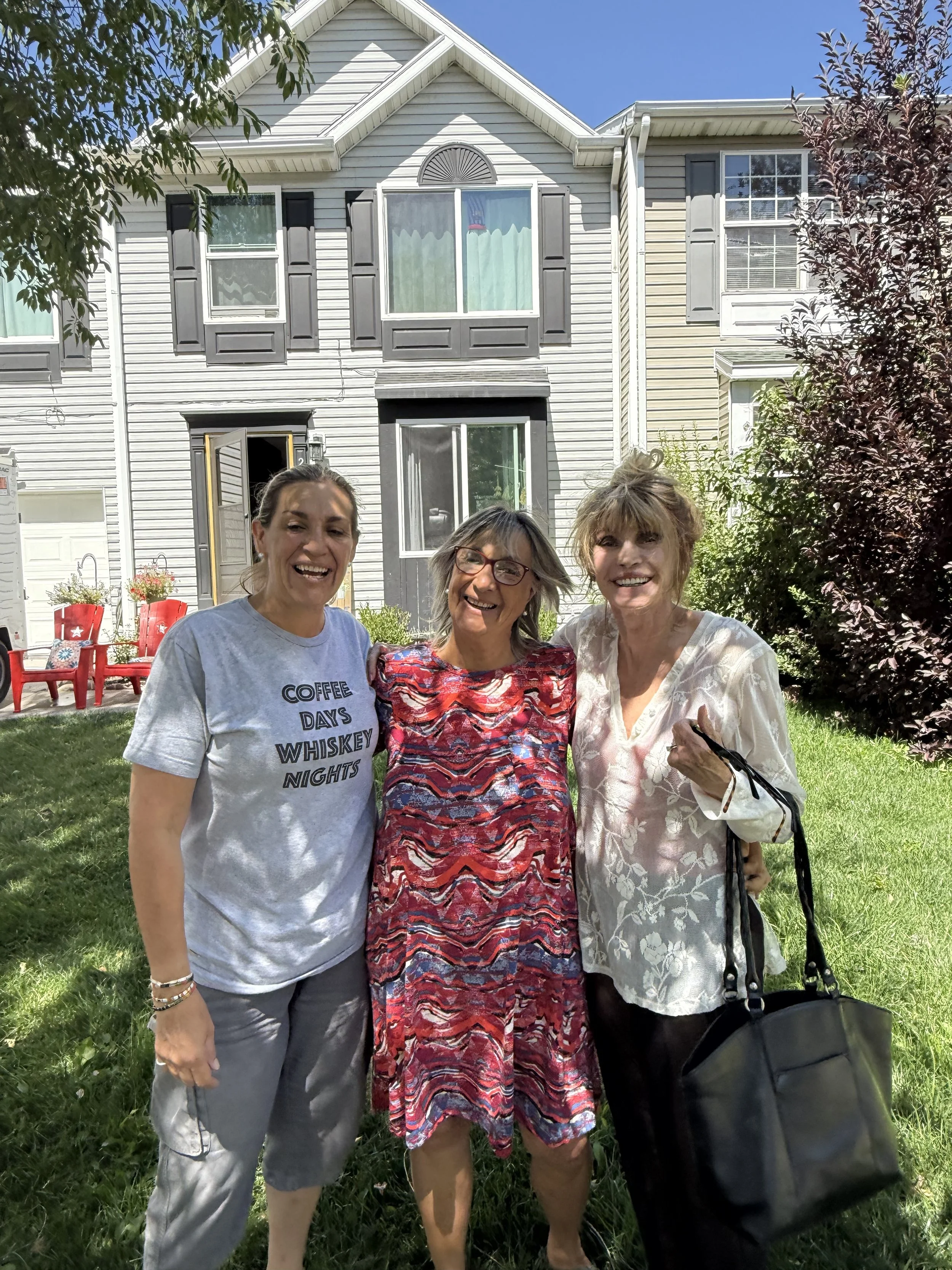 Three women standing outside on a grassy lawn in front of a silver multi-story house with black shutters, smiling and posing for the photo on a sunny day.