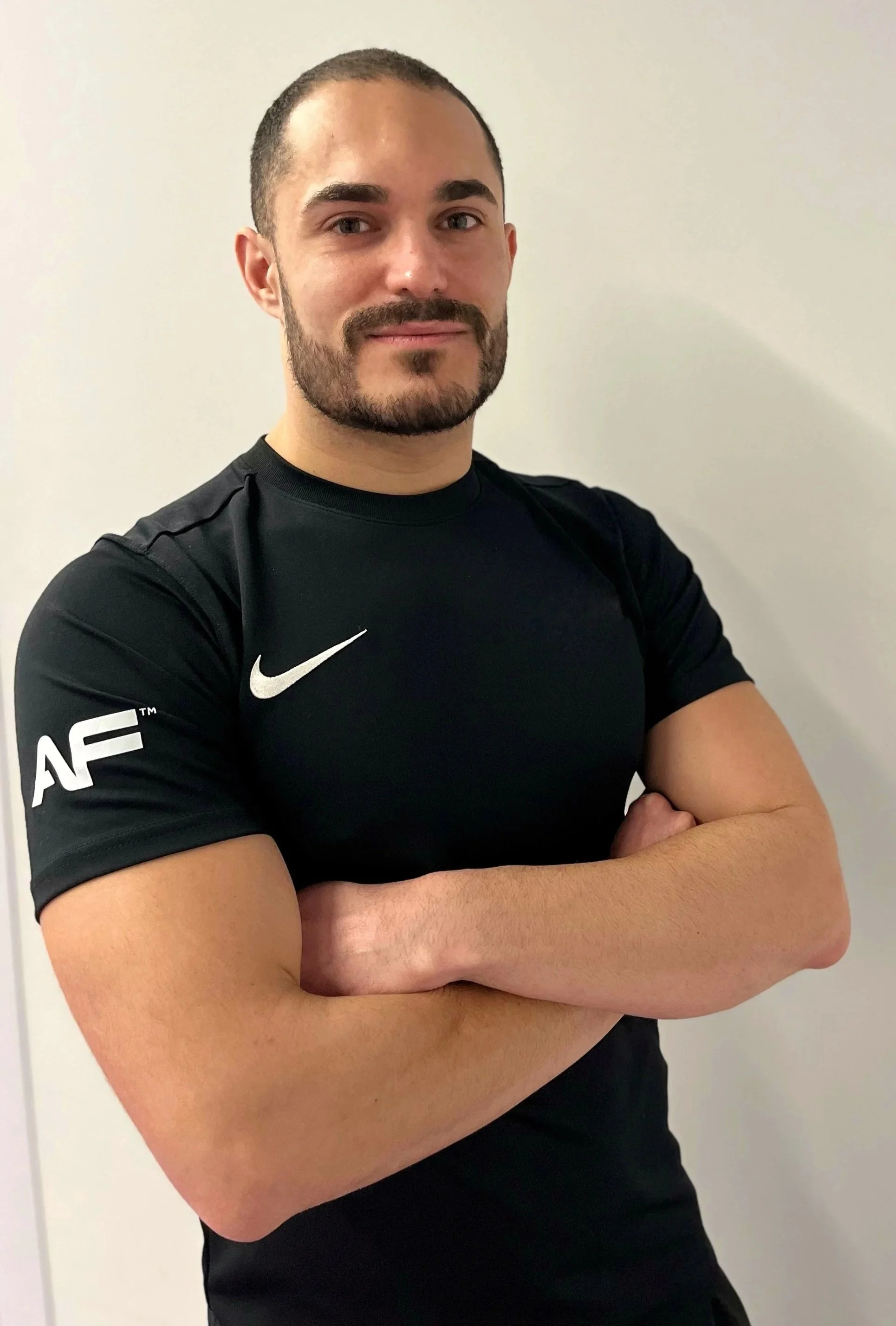 Portrait of a man with short dark hair and a beard, wearing a black Nike sports shirt with white logo, standing against a plain light-colored wall with arms crossed.