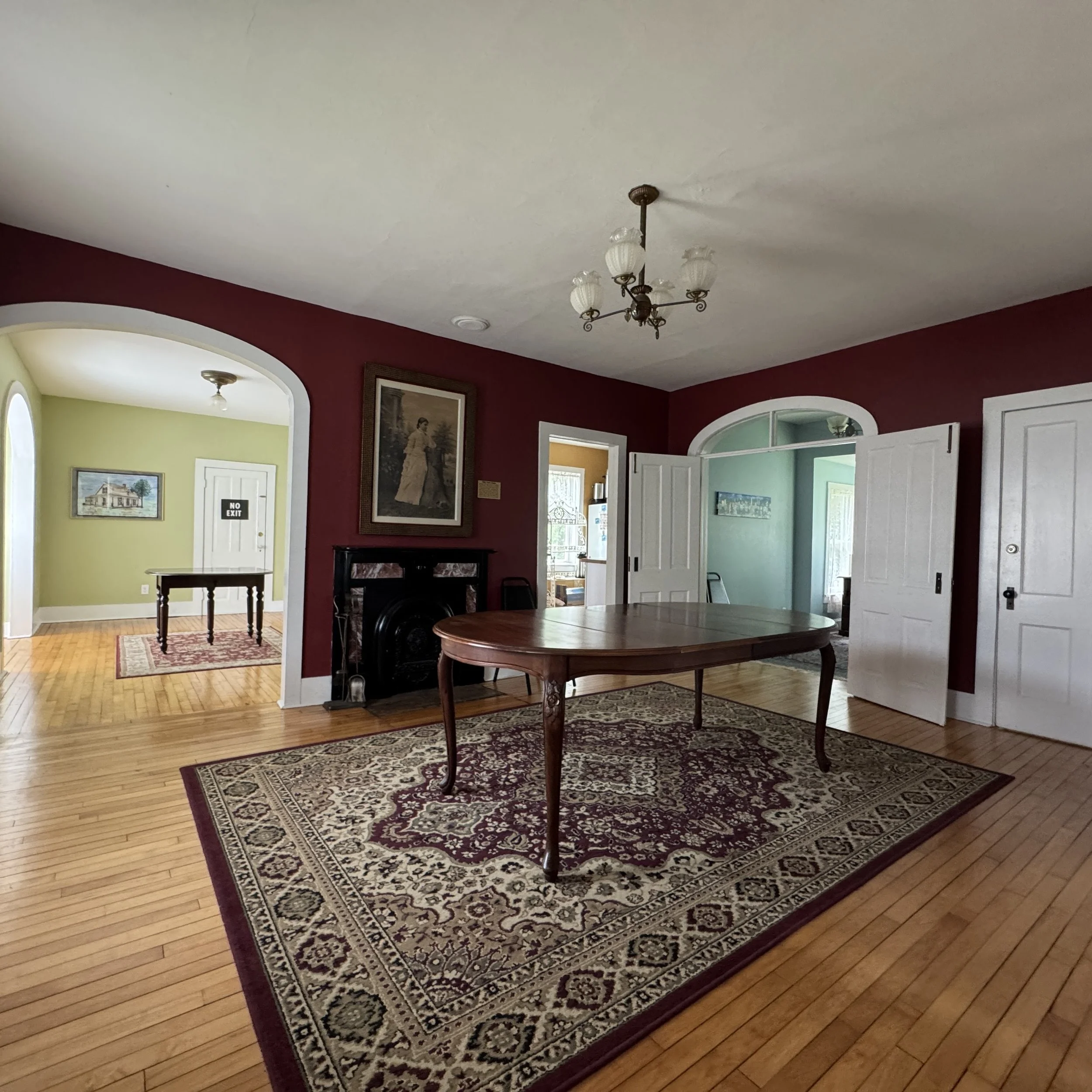 Living room with a wooden table on a patterned area rug, red walls, a chandelier, a fireplace, and open doorways leading to other rooms.