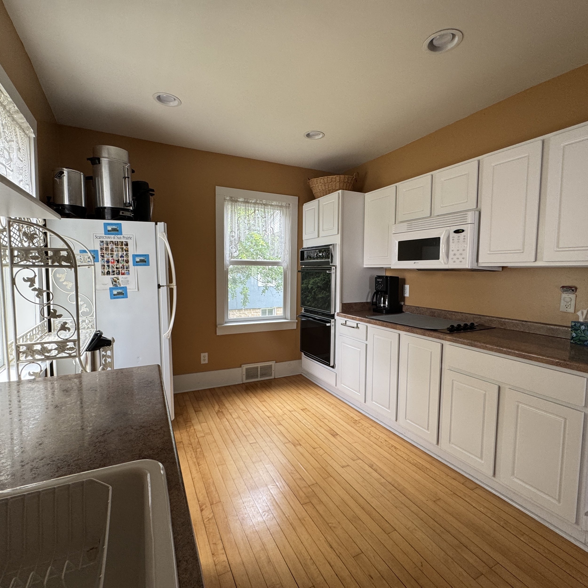 Kitchen with hardwood floor, white cabinets, beige walls, window with lace curtains, white refrigerator, microwave, coffee maker, stove, and sink.