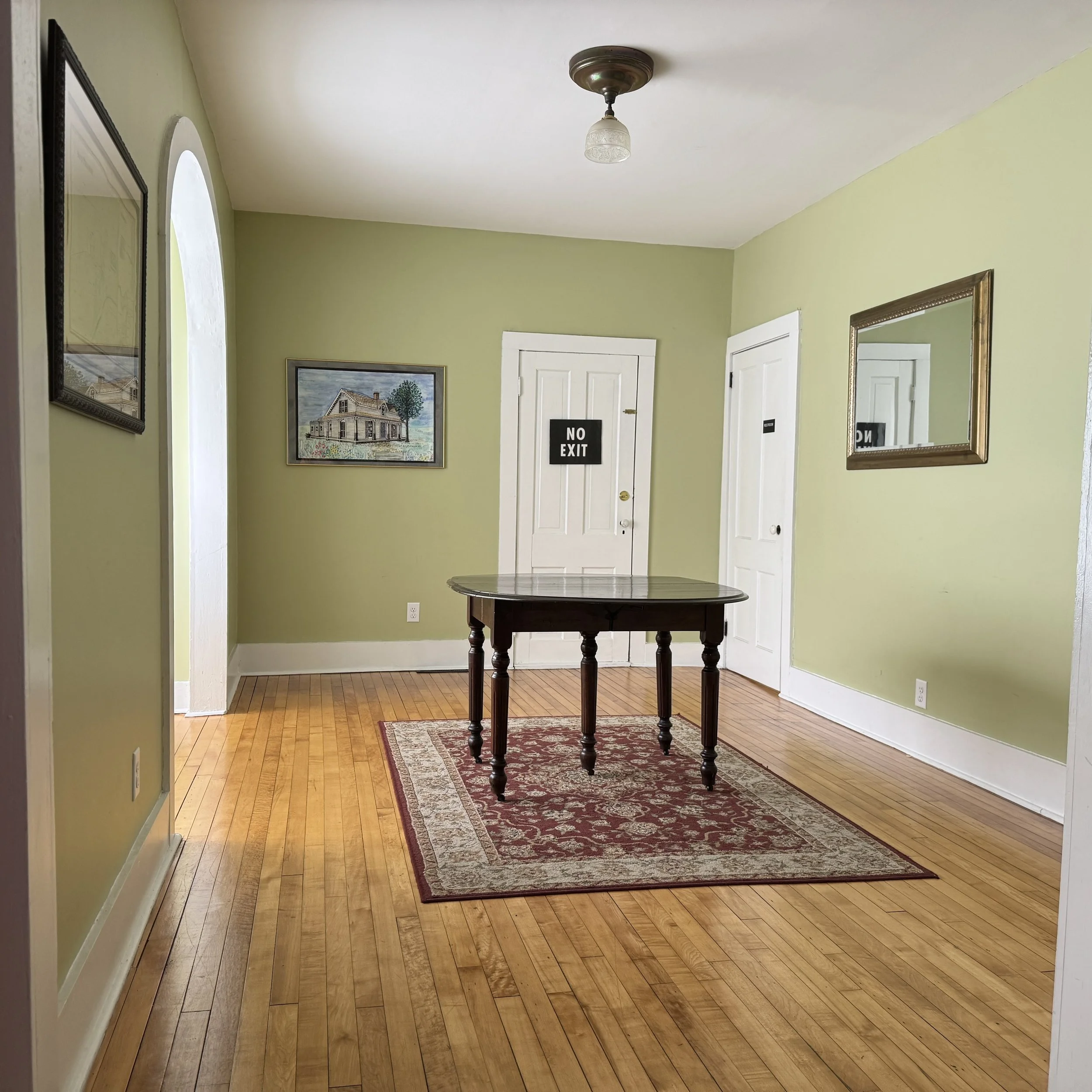 Empty room with green walls, hardwood floor, a small rug, a dark wooden table, and two white doors, one with a 'NO EXIT' sign, and framed pictures on the walls.